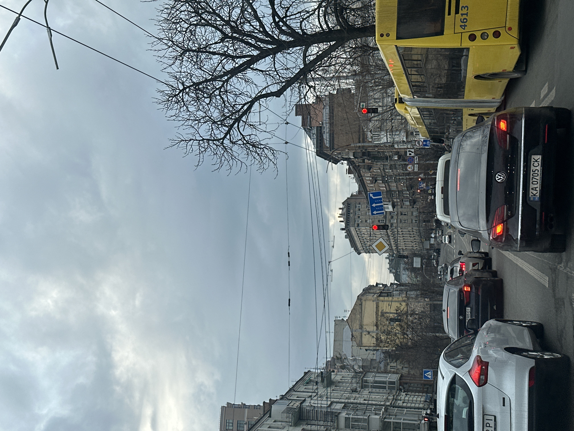 The image depicts a street scene in Kyiv, Ukraine. The view is oriented along the street, looking towards a row of buildings on either side. Traffic is visible in the foreground, including a yellow bus and several cars, one of which has a license plate reading "KA 0705 CK". Traffic lights can be seen, some of which are red. Power lines span across the sky. A bare tree is visible on the left side of the image. Buildings of varying heights and architectural styles line the street. The sky is overcast.
