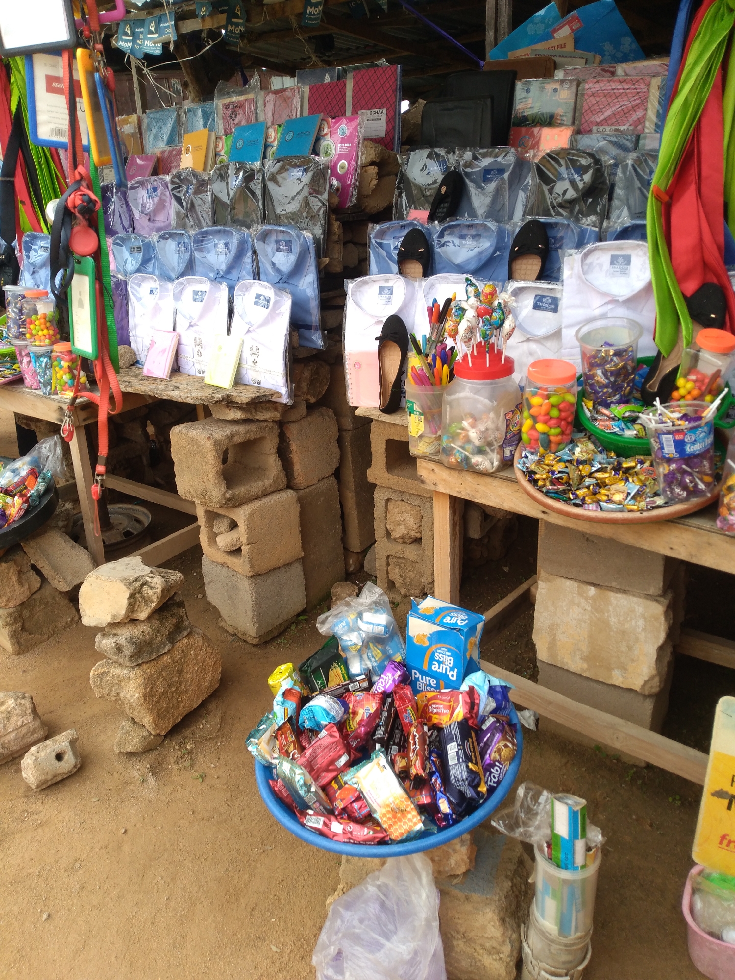 This is a wide-angle, eye-level outdoor shot of a makeshift stall in Jos, Nigeria, displaying a variety of goods for sale. The stall appears to be set up along a dusty, uneven ground, with concrete blocks used as makeshift supports and display structures.
In the foreground, a blue plastic basin is filled with an assortment of packaged snacks and candies, including items with "Pure Bliss" branding. To the right of the basin, a tall cylindrical container holds more packaged snacks. Further right, a pink bucket contains some items, partially obscured.
The middle ground features a wooden table supported by more concrete blocks. On this table, various items are displayed, including jars of colorful candies, a tray of individually wrapped candies, and lollipops arranged in a red container. Several pairs of women's shoes, mostly black, are also visible, some placed on the table and others on shelves above. Shelves above the table are stocked with neatly folded shirts, predominantly white, blue, and light purple, each individually packaged in plastic. Behind the shirts, more packaged goods, possibly stationery or notebooks, in various colors like pink, blue, and yellow, are stacked.
The background shows a corrugated metal roof providing shelter for the stall. Various hanging items, including colorful straps, lanyards, and fabric rolls (red, green, and yellow), are suspended from the roof structure. Some blue banners with text are also visible hanging from the roof. The overall impression is that of a busy, informal market stall catering to everyday needs in an urban environment.