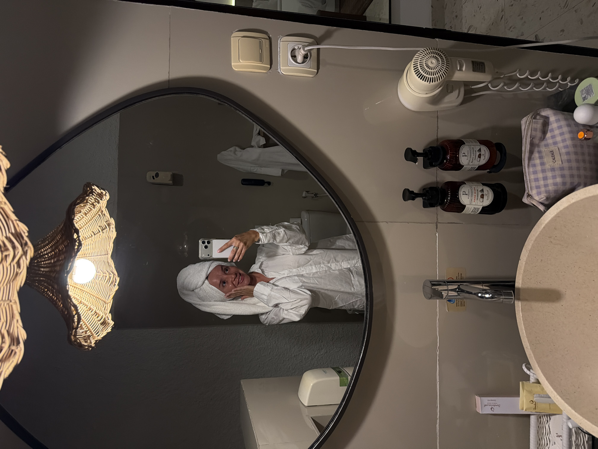 A woman with a towel wrapped around her head and wearing a white button-down shirt is reflected in a large oval mirror in a bathroom. She is holding her phone to take a selfie, with her hands touching her face. To the left of the mirror, a white soap dispenser is mounted on the wall. In the reflection, a white bathrobe is hanging on a hook. Below the mirror, a beige sink is visible, with a chrome faucet. To the right of the sink, two bottles of hand soap and body lotion are mounted on the wall, along with a white hairdryer. A checkered toiletry bag is placed on the counter in front of the bottles. The bathroom appears to be modern and clean, with neutral-colored walls and tiled surfaces. The overall lighting is warm, likely from the woven pendant lamp above the mirror, casting a soft glow on the scene. The location is identified as Unknown, Indonesia.