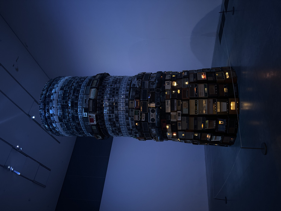 A tall, cylindrical art installation, approximately four to five meters high, dominates the center of a dimly lit exhibition hall. The installation is constructed from hundreds of stacked vintage radios, boomboxes, and other audio components, forming a tapering tower. These devices vary in size, color, and design, with some showing illuminated dials or screens, casting warm orange and white glows, particularly evident in the lower sections of the tower. The surrounding environment consists of plain, light-colored walls bathed in a deep blue ambient light, a dark gray concrete floor, and a high ceiling with exposed track lighting fixtures. A white rope barrier, supported by metal stanchions, encircles the base of the artwork, indicating a designated viewing distance. No individuals are present in the scene. The location is the Tate Modern, an art gallery situated in London, United Kingdom.