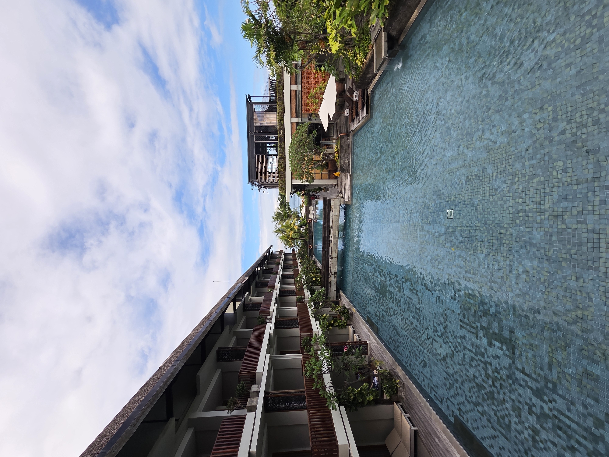 The image shows a view in North Kuta, Indonesia. The perspective is unusual, looking vertically up between a building on the left and a swimming pool on the right, with the sky overhead. The building appears to be a multi-story hotel with balconies adorned with plants. The swimming pool is long and narrow, with tiled walls. The sky is blue with scattered white clouds. On the far side of the pool, there's another building with outdoor seating and greenery. There are no people visible in the scene.