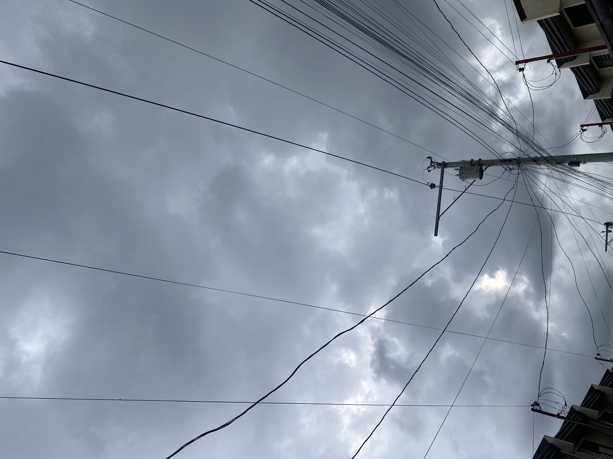 The image depicts a low-angle shot of a sky filled with dark, overcast clouds, suggesting impending rain. Several overhead electrical wires crisscross the frame, creating a complex, tangled network. A utility pole with a transformer and numerous wires attached stands prominently in the mid-right of the image. In the lower portion of the frame, the rooftops of buildings, appearing to be houses or townhouses, are visible on the left and right edges. The architectural style and the presence of such utility infrastructure are consistent with residential areas in Davao City, Philippines. No people are visible in the frame. The overall atmosphere is somber due to the heavy cloud cover and the dense arrangement of electrical wires.