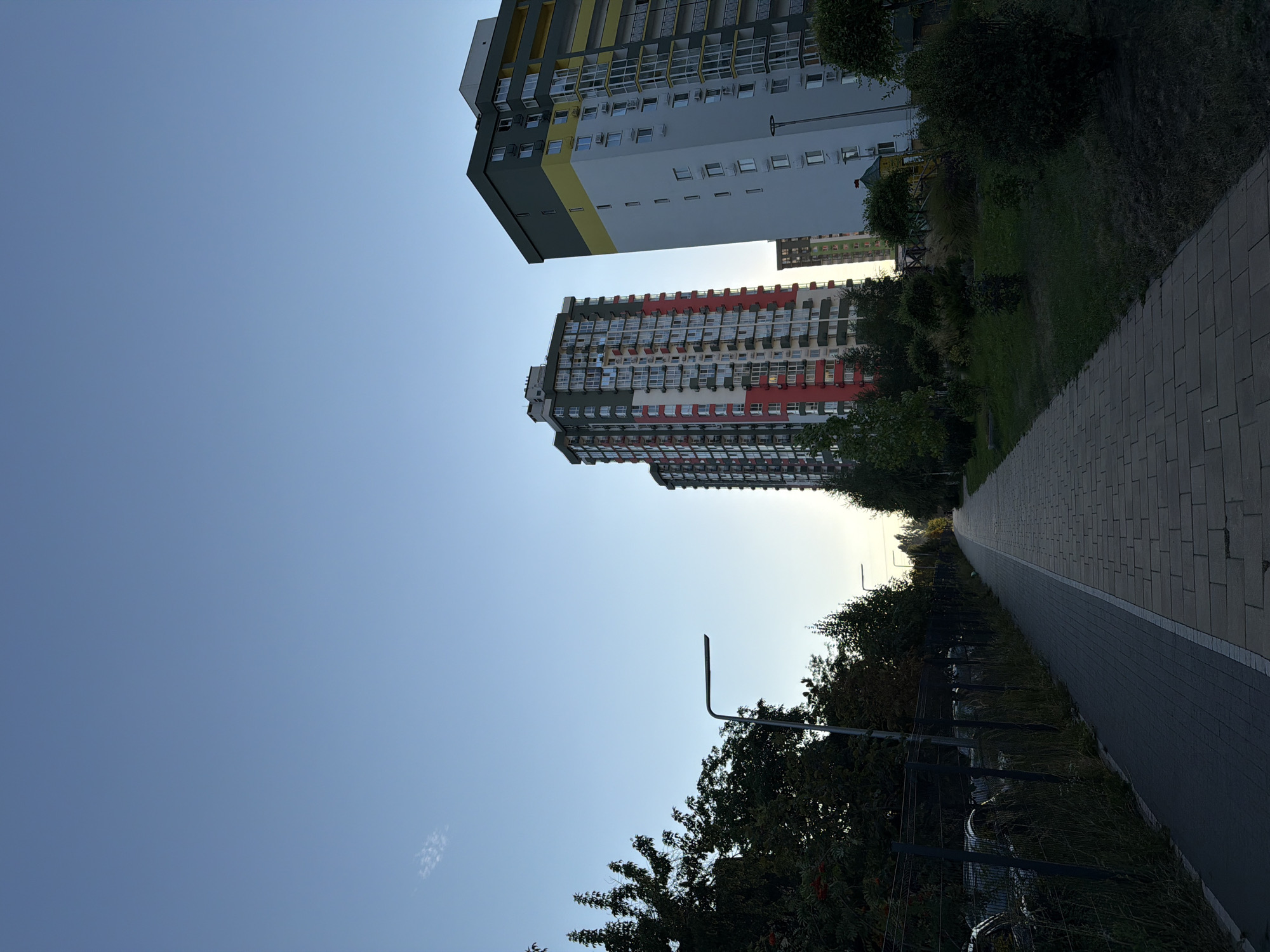 This is a ground-level, vertical, outdoor photograph taken during the daytime, depicting a modern residential area in Kyiv, Ukraine. The composition is dominated by tall, multi-story apartment buildings with contemporary architectural designs. The building on the left features a facade with alternating sections of red, white, and grey, punctuated by numerous windows and balconies. To its right stands another apartment building, taller and wider, with a distinctive color scheme of grey at the top, a broad yellow band, and white lower sections. The buildings are situated in a newly developed urban landscape.
In the foreground, a paved walkway, constructed with rectangular grey bricks, stretches into the distance, flanked by a darker, narrower path on the left, possibly a bicycle lane or sidewalk. Adjacent to the darker path is a dark metal fence, behind which parked cars are visible. Lush green trees and shrubs are scattered throughout the scene, with denser foliage lining the left side of the walkway and smaller bushes and manicured greenery to the right. A street lamp with a modern, minimalist design is positioned along the left side of the walkway.
The sky is a clear, soft blue, with a hint of lighter blue near the horizon, suggesting it is either early morning or late afternoon. A faint, wispy cloud is visible in the upper left quadrant of the image. The overall atmosphere is one of urban development and residential comfort. There are no people visible in the frame.