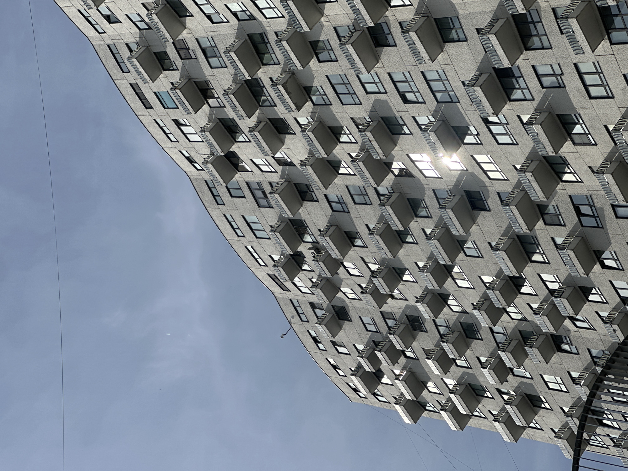 The image depicts a multi-story building in Moscow, Russia, with a facade characterized by numerous projecting balconies. The building's exterior is light gray, and the balconies cast shadows on the facade. The sky is a pale blue with some cloud cover. A thin power line is visible extending across the sky.