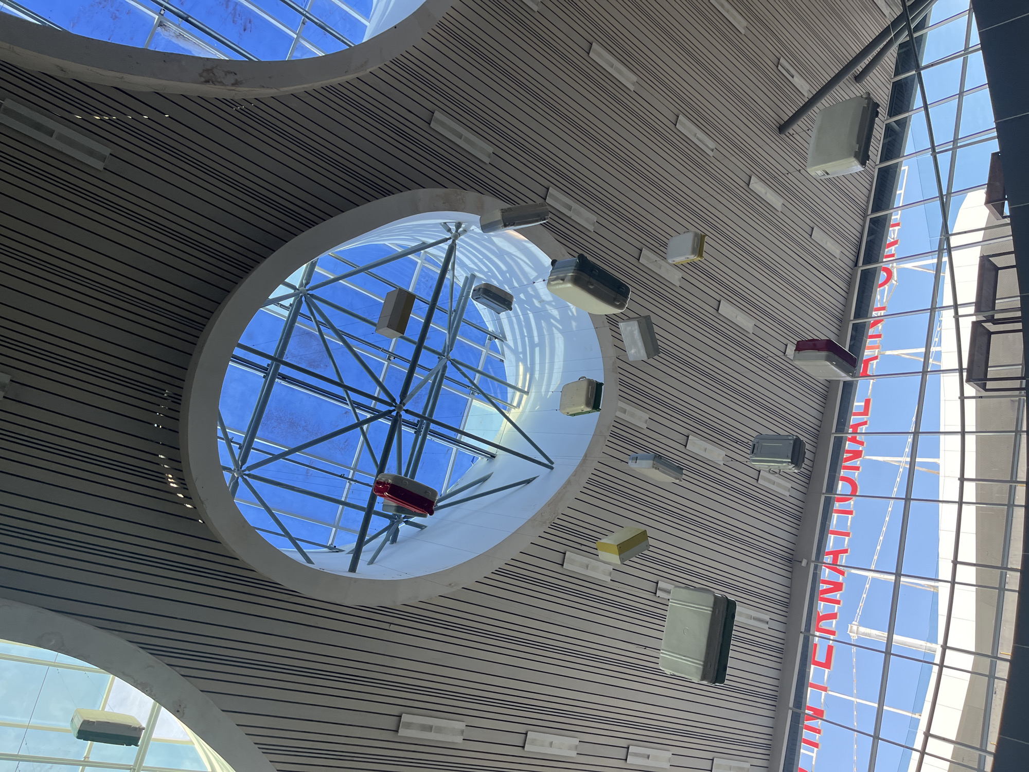 The image depicts an interior view of Kuala Namu International Airport (KNO) in Ramunia Dua, Indonesia. The ceiling is characterized by horizontal stripes and circular openings revealing a blue sky and structural supports. Numerous suitcases are suspended from the ceiling at various points. A glass facade with red lettering spells out "INTERNATIONAL".