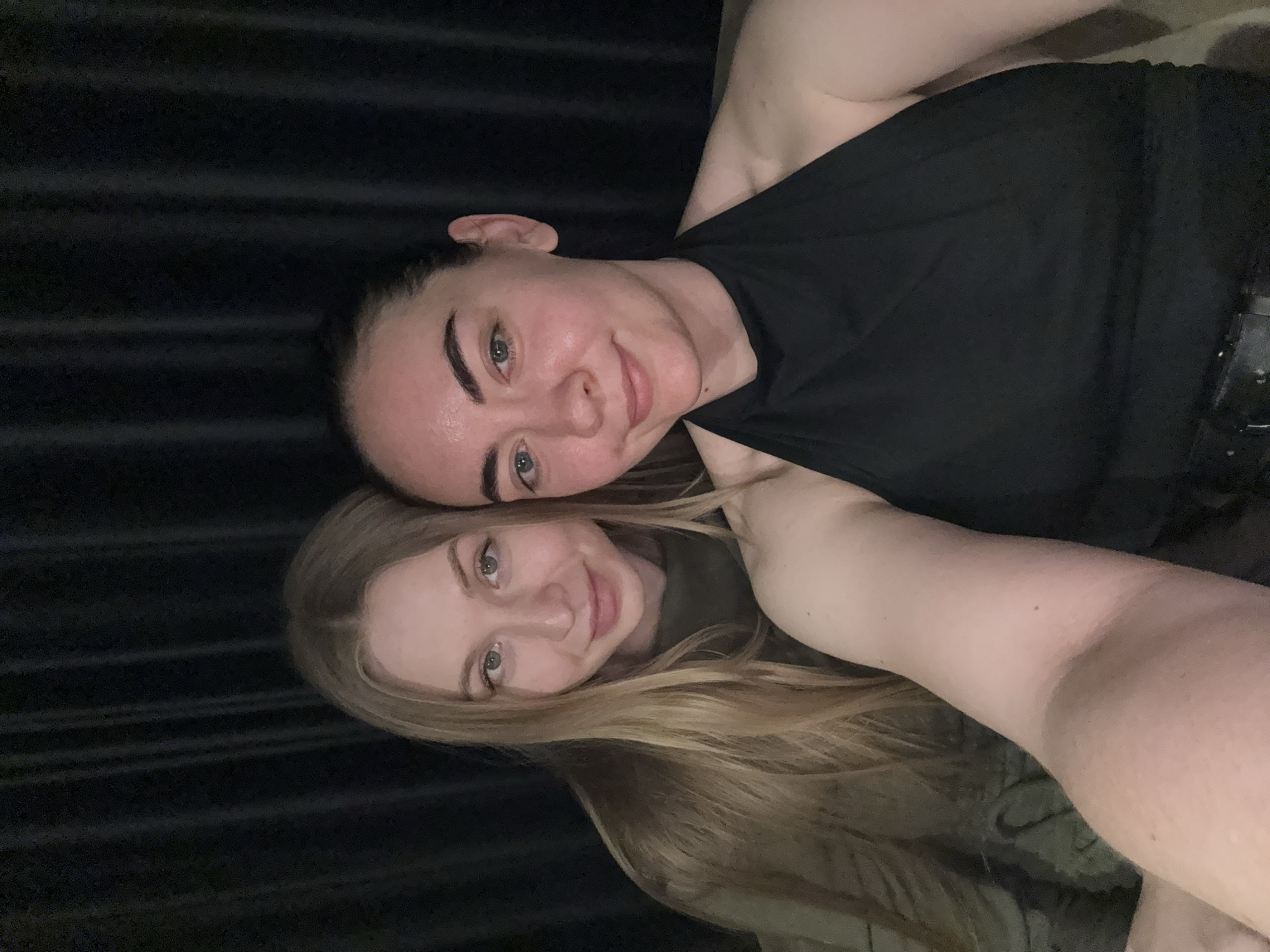 Two women are posed together indoors. The woman closer to the top of the frame has dark hair pulled back, and is wearing a black tank top. The woman below has long, blond hair, and is wearing a dark green top. The background is dark. No information indicates this image was taken in Chaikovsky, Russia.