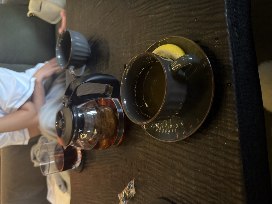 The scene depicts an indoor dining or lounge area located in Tibubeneng, Indonesia. A dark wooden table serves as the primary surface. In the foreground, a dark gray ceramic teacup containing a light-colored liquid rests on a matching dark gray saucer with a faint decorative pattern; a yellow lemon wedge is positioned on the saucer. Centrally, a clear glass teapot with a black handle and lid holds amber-colored tea with an internal metal infuser. To the left of the teapot, a clear glass tumbler containing a dark red beverage sits on a dark coaster. Behind the teapot, an empty, ridged dark gray ceramic teacup is partially visible. A small, crinkled silver foil packet lies on the table to the left. In the background, an individual is partially visible, seated on a dark-colored sofa, wearing a white top with light blue trim and light-colored lower garments. A light-colored bag and a folded white textile are also present on the sofa. No direct actions or interactions between individuals are visible.