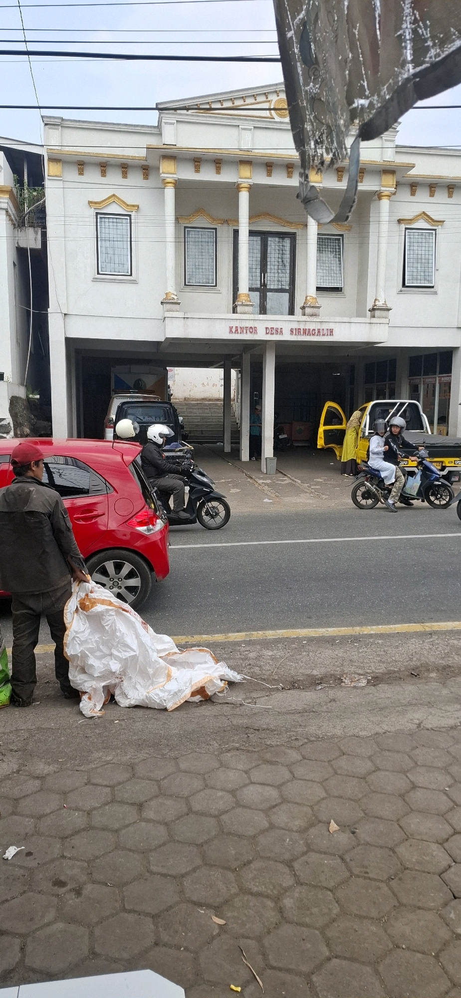 In front of the Sirnagalih village office in Sirnagalih, Indonesia, a man wearing a red cap and dark clothing stands on a hexagonal-tiled sidewalk, holding a large, crumpled white object with orange trim. To his left, a red car is parked, and to his right, a motorcycle with two riders wearing white helmets and masks passes by on the asphalt road. Behind the red car, a black motorcycle with a single rider in a black jacket and white helmet is also on the road. The village office is a two-story building with white walls, gold decorative columns, and multiple windows. A yellow pickup truck with its door open is parked under the covered area of the building. The sky is overcast.