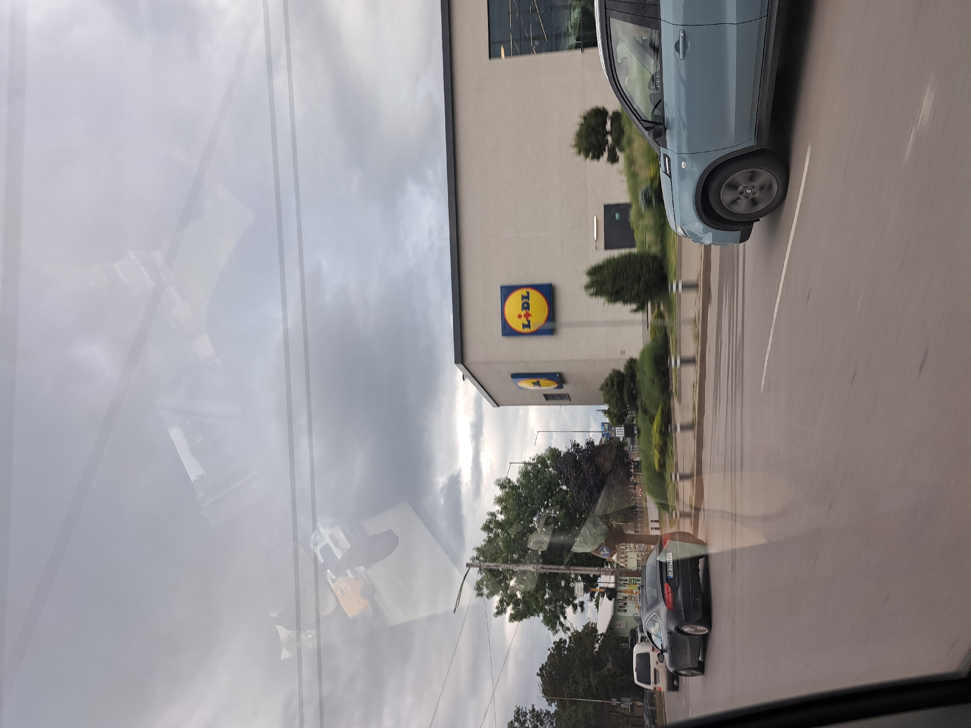 The image shows a view from inside a car in Sofia, Bulgaria, driving past a Lidl store. The Lidl logo is prominently displayed on the building. Another car is visible in the opposite lane. The sky is cloudy. The image is taken through the car's windshield, which shows some reflection of the sky and surroundings.