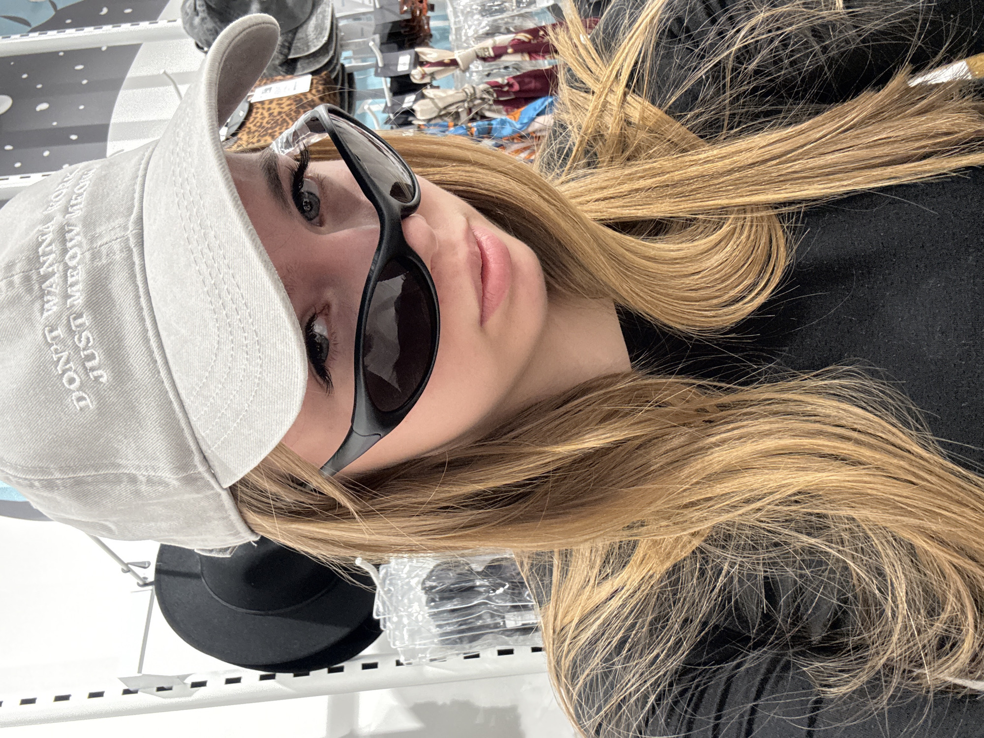 A headshot-style image depicts a young female inside an accessories store in Sochi, Russia. The subject is centrally framed, looking directly at the camera with a neutral expression. She wears a light grey baseball cap with white embroidered text visible on the front, partially reading "DON'T WANNA" and "JUST MEOW MEOW". Dark, wrap-around style sunglasses are positioned low on her nose, allowing her grey-blue eyes and prominent eyelashes to be seen above the frames. Her long, straight, blonde hair is parted in the middle and extends over her shoulders, covering parts of her black long-sleeved top. The background, largely blurred, indicates an indoor retail environment with white shelving units. Visible items on display include a dark circular hat, various packaged accessories in clear plastic, a leopard-print item, and multiple patterned scarves or hair accessories on hooks.