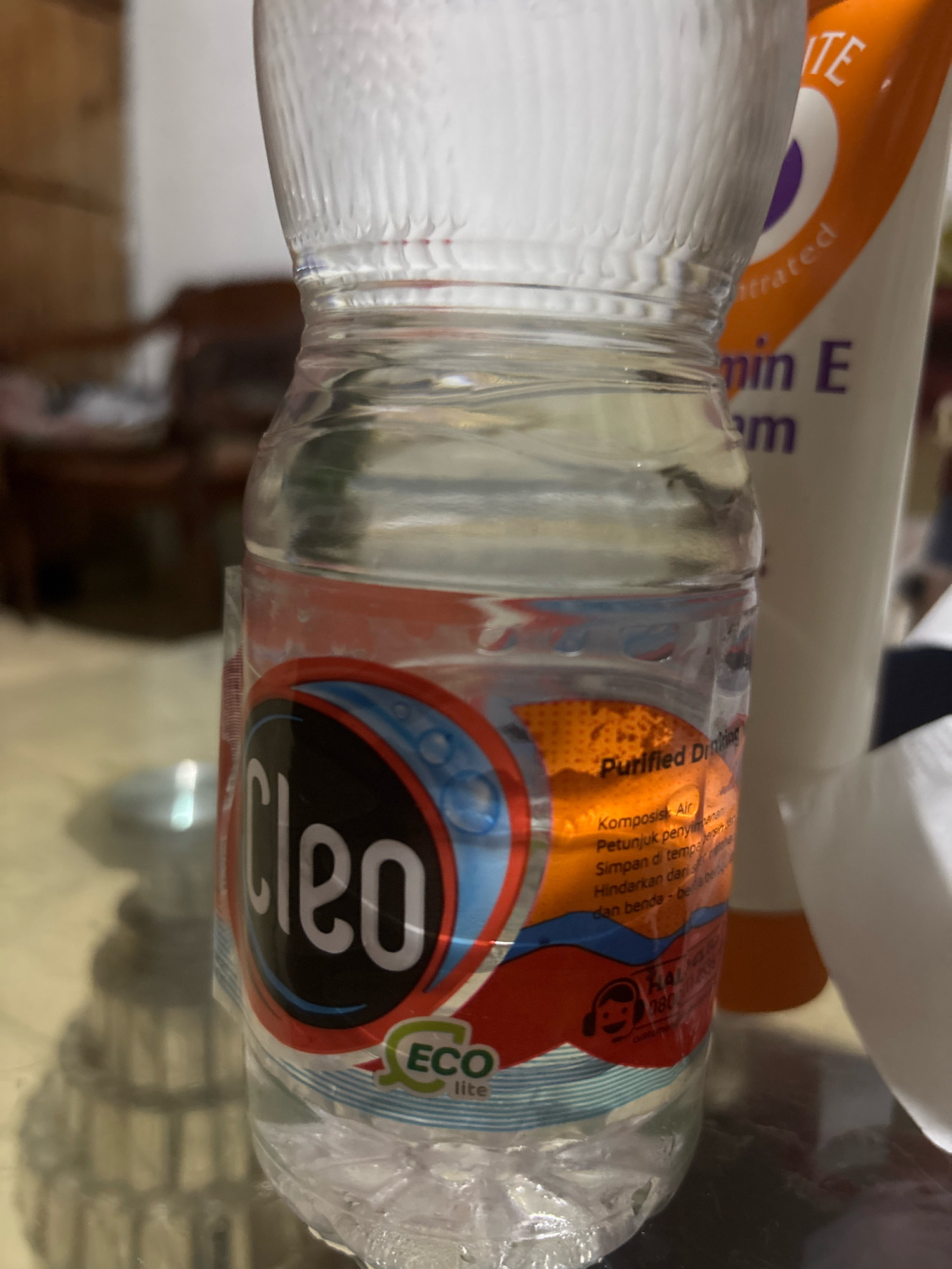 A close-up, indoor shot captures a clear plastic bottle of purified drinking water, labeled "Cleo Eco Lite," positioned on a reflective surface that appears to be a table. The bottle's label is predominantly red, white, and blue, featuring the brand name "Cleo" in a distinctive black font. A partially visible white and orange tube, likely a cosmetic product, stands behind the water bottle. The background is blurred, suggesting an interior setting with indistinct furniture. The lighting is moderate, casting subtle shadows. The image is taken from a slightly elevated angle, focusing on the water bottle. No specific city or location in Bekasi, Indonesia, is identifiable from the visual information presented.