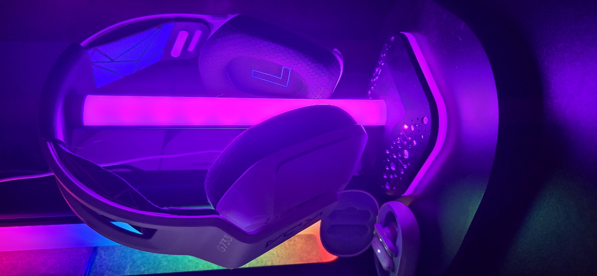 The image captures a pair of white Logitech G733 gaming headphones displayed in an indoor setting, likely a gaming setup or desk, under vibrant LED lighting. The headphones are oriented with one earcup facing the viewer, clearly showing the text "G733" on its side. A subtle 'L' shape design is visible on the headband.
A prominent pink/magenta LED light strip horizontally illuminates the area directly behind the headphones, casting strong purple and pink reflections throughout the scene. Further down, other LED lights in blue, green, and orange/red contribute to a multicolored, atmospheric glow. The overall environment is dark, suggesting the photograph was taken at night or in a dimly lit room, with the artificial lighting being the dominant light source.
To the right of the headphones, part of the stand or a piece of equipment with a perforated, dotted pattern is visible. Below the headphones, an open charging case containing two wireless earbuds is also present, bathed in the colorful light. The scene creates a modern, tech-focused aesthetic. The photograph was taken in Burgas, Bulgaria.