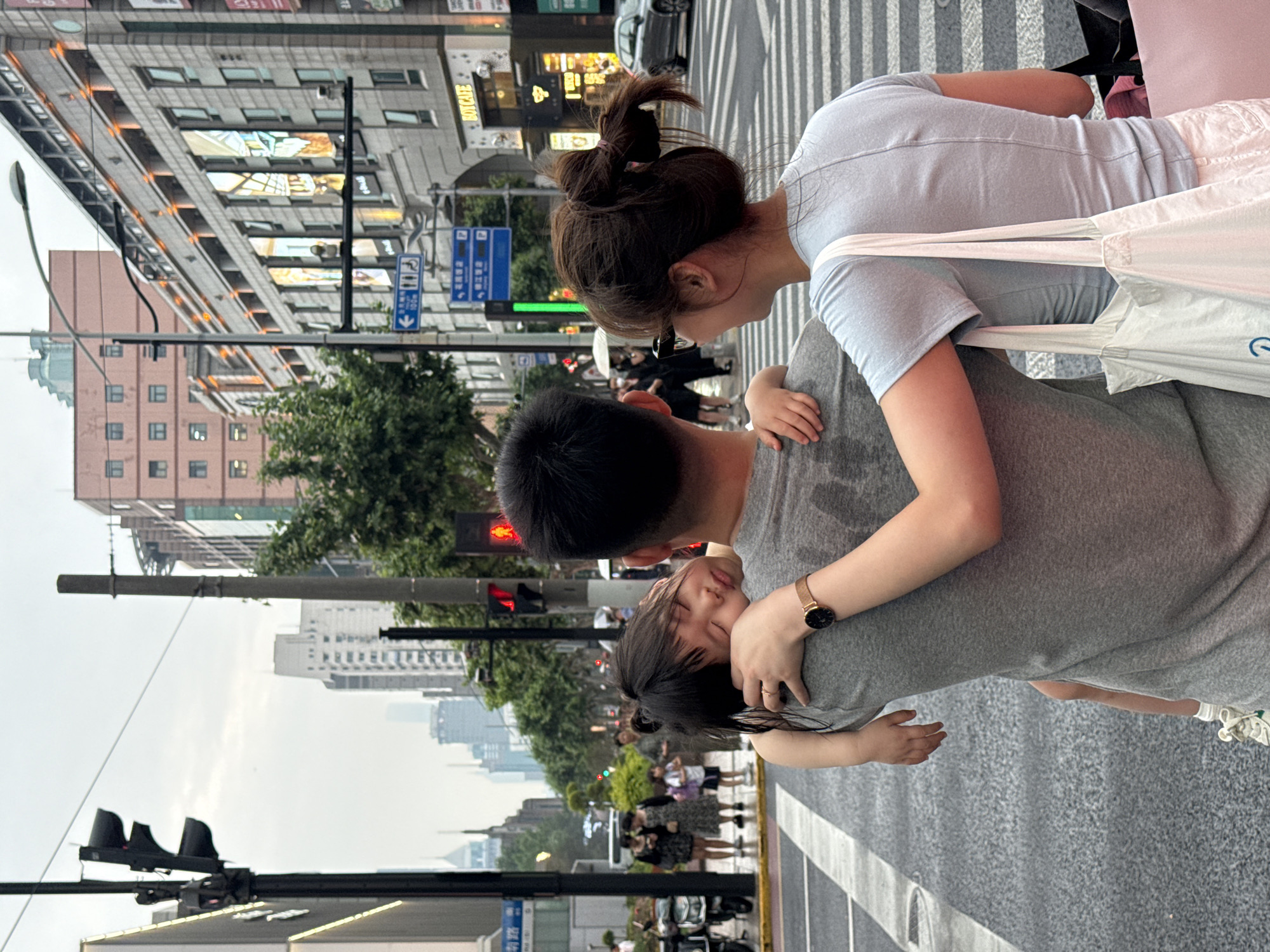 An urban street scene, rotated 90 degrees counter-clockwise, depicts a family on what appears to be a wet road or sidewalk, suggesting recent rain. In the foreground, a man in a grey t-shirt is seen from behind/side, carrying a young child who is nestled and seemingly asleep or resting on his shoulder. The child has dark hair and a small hand is visible. Next to them, a woman, also seen from behind/side, wears a light blue t-shirt, white shorts, and has dark hair tied in a bun. She carries a white shoulder bag on her left side and wears a watch with a gold strap and dark face on her left wrist.
The setting is a bustling city street, identified as Xuhui District, China. In the background, tall, multi-story buildings line the street, some with illuminated windows or signs. One prominent building sign in the mid-ground clearly reads "BOYCAFE" in English, alongside Chinese characters. Traffic infrastructure includes zebra-striped crosswalks, traffic lights (one showing red), street lamps, and blue road signs with white Chinese characters and symbols, including a wheelchair accessibility symbol and "100m." Other pedestrians are visible in the blurred background. The sky is overcast, and the overall lighting suggests either a heavily cloudy day or late afternoon/early evening. The image captures a tender, candid moment of parental care amidst the urban environment.
