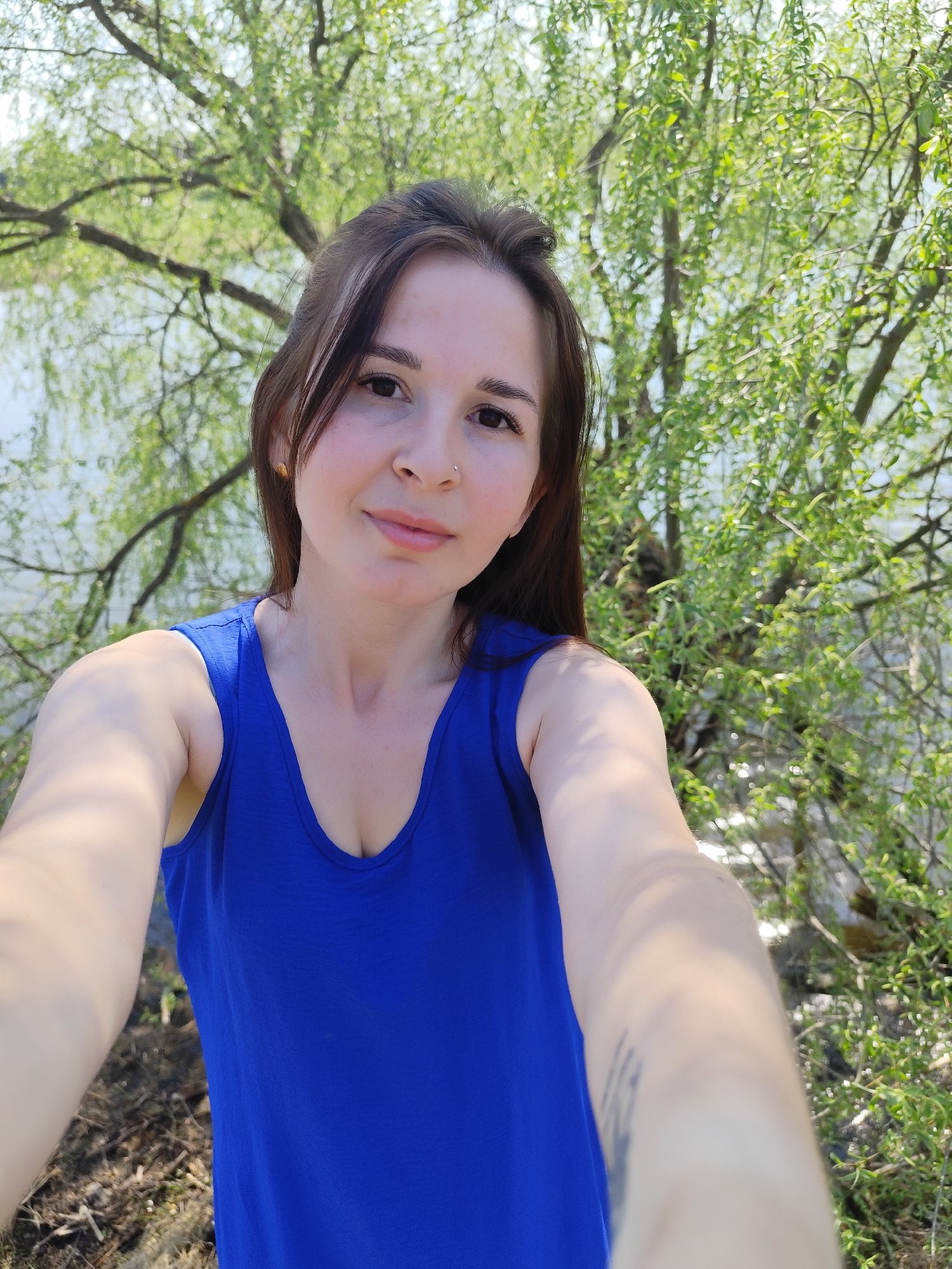 A woman is taking a selfie outdoors. She has brown hair and is wearing a blue tank top. Behind her is a body of water and a tree with green leaves. The location is likely in or around Фоки, Russia, based on the prompt.