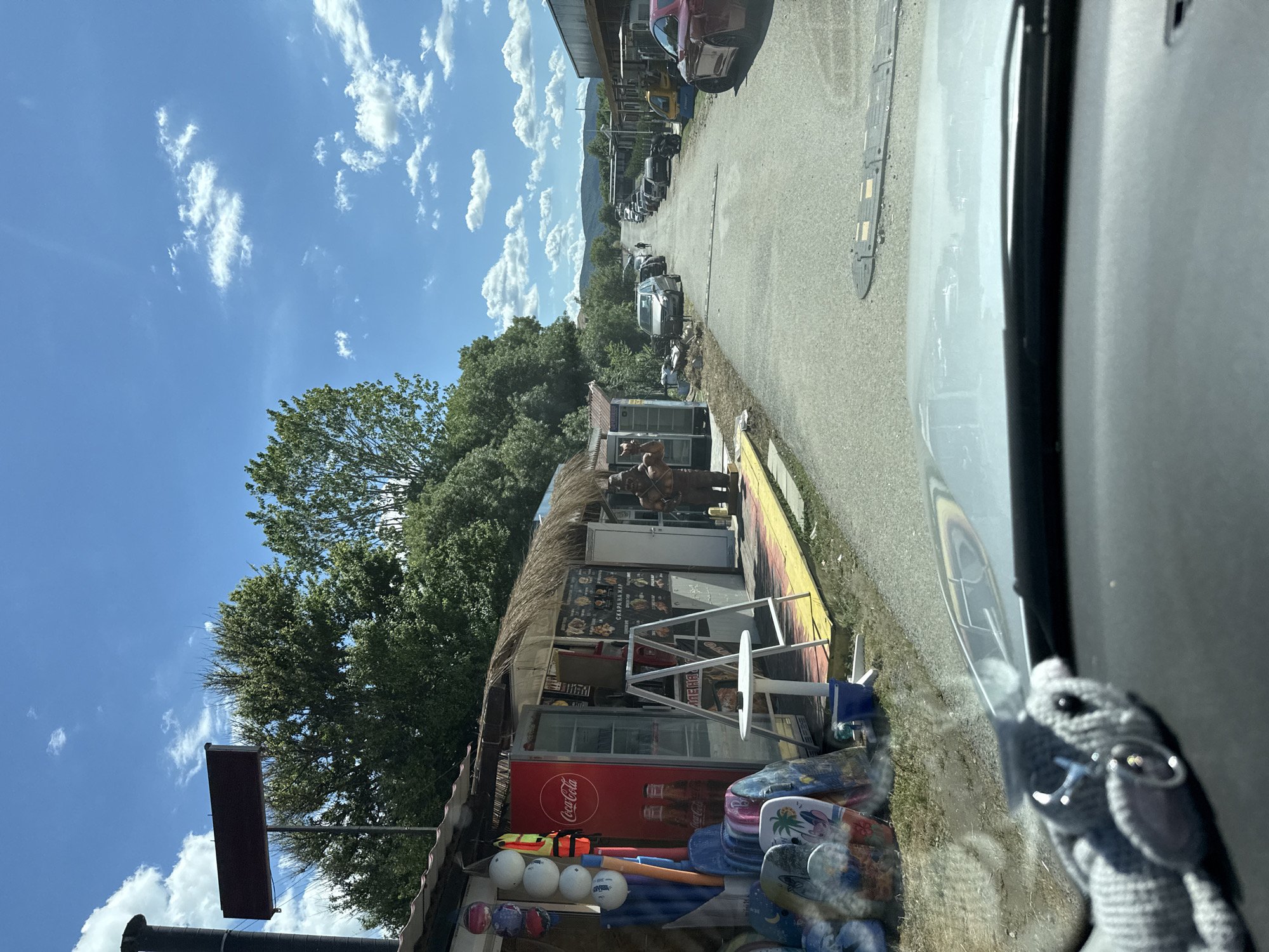 A scene captured from inside a vehicle shows a small road lined with buildings and shops, likely a commercial area in Tsarevo, Bulgaria. The shops on the left appear to be casual outdoor stalls, selling items like surfboards, life vests, and refreshments from a Coca-Cola branded cooler. One stall has a thatched roof and a menu board displaying food items. A life-sized statue of what appears to be a character in armor stands outside one of the shops. The road itself is paved and has speed bumps. Several cars are parked along the road, and a few are driving away in the distance. The environment suggests a sunny day with blue skies and scattered clouds. The foreground shows the interior of the vehicle, including a dashboard and a stuffed animal hanging from the rearview mirror.