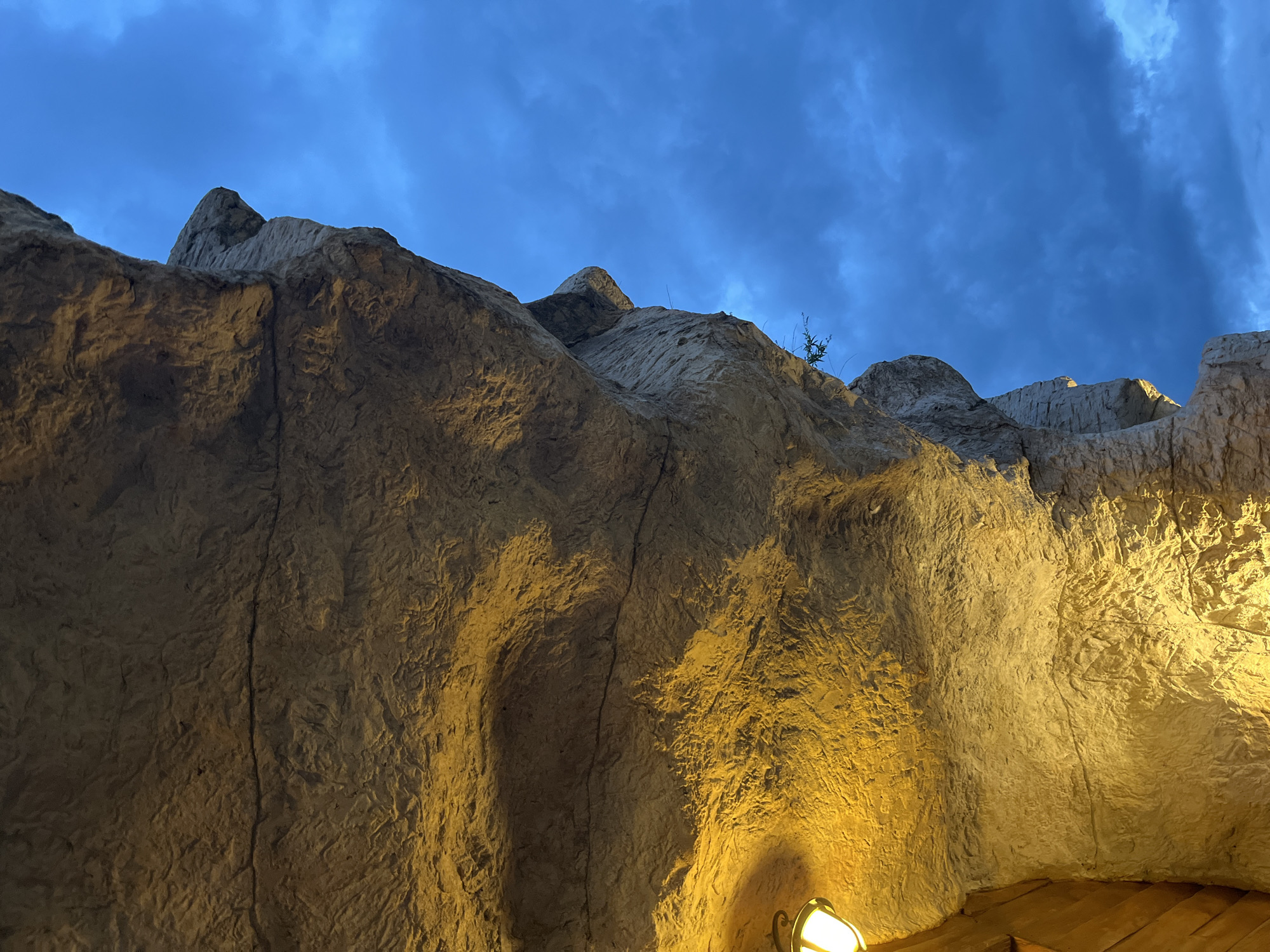 The image shows a low-angle view of a rock formation against a twilight sky, likely artificial as it is indoors. The sky is dark blue with visible clouds. The rock formation occupies most of the frame and exhibits a rough, textured surface with a light tan/beige color, illuminated by artificial light emanating from below, indicating its location is in the city of Koprivlen, Bulgaria. A light fixture and wooden structure are visible at the bottom right, suggesting this is an indoor artificial environment. No people are visible. The overall scene is static and emphasizes the architectural design of the rock structure.