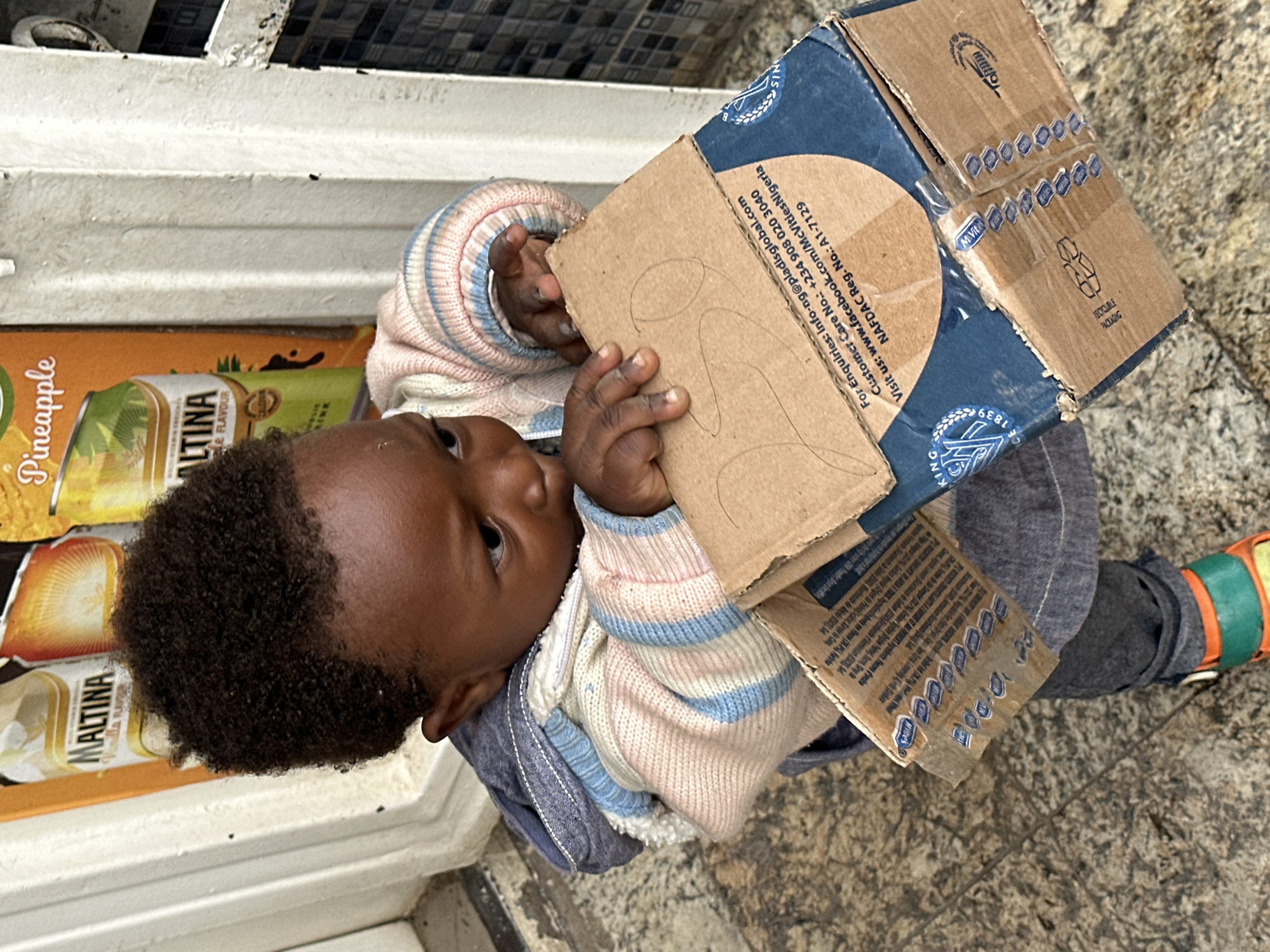 A young child, likely a boy, is crouched down, holding a cardboard box in front of him. He is wearing a light-colored sweater with horizontal stripes of pink, light blue, and white, over a dark grey hooded jacket and pants. His right foot is visible, wearing a colorful sandal with orange and green straps. The child's eyes are looking downwards at the box, and his hands are gripping its edges.
The box appears to be a product package, with some text and logos visible on its surface. The text includes an email address "Info-ng@pladisglobal.com", a phone number "+234 908 020 3040", and a website address "www.facebook.com/McVitiesNigeria". There is also a NAFDAC Registration number "A1-7129". A circular blue logo with the letters "McV" and "1839" is prominent. Hand-drawn numbers "20" are visible on the top surface of the box.
The background shows a wall with a white framed window or doorway, and a tiled section on the right. In the upper left, a poster with images of canned drinks, including "MALTINA Vanilla Flavour" and "Pineapple", is partially visible. The ground is paved with speckled grey tiles.
The scene is likely an outdoor setting, possibly in front of a shop or market stall, consistent with a location in Gura Top, Nigeria. The lighting suggests daylight.