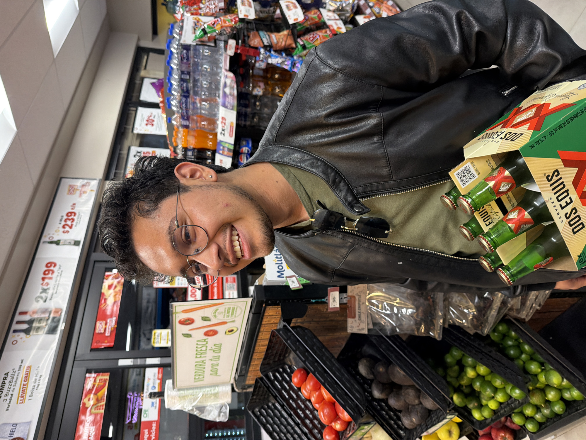 The image depicts a man in a store, possibly a convenience store or a small supermarket in San Andrés Cholula, Mexico. He has short, dark curly hair, wears glasses and a black leather jacket over a green shirt. He is smiling and holding a case of "Dos Equis" beer. In the foreground, there are several black plastic baskets filled with produce, including tomatoes, avocados, and limes. In the background, shelves are visible, stocked with various products like bottled drinks and snacks. Price tags in the local currency (pesos) are visible on the shelves, and a sign promoting "Verdura Fresca" (Fresh Vegetables) is also visible.