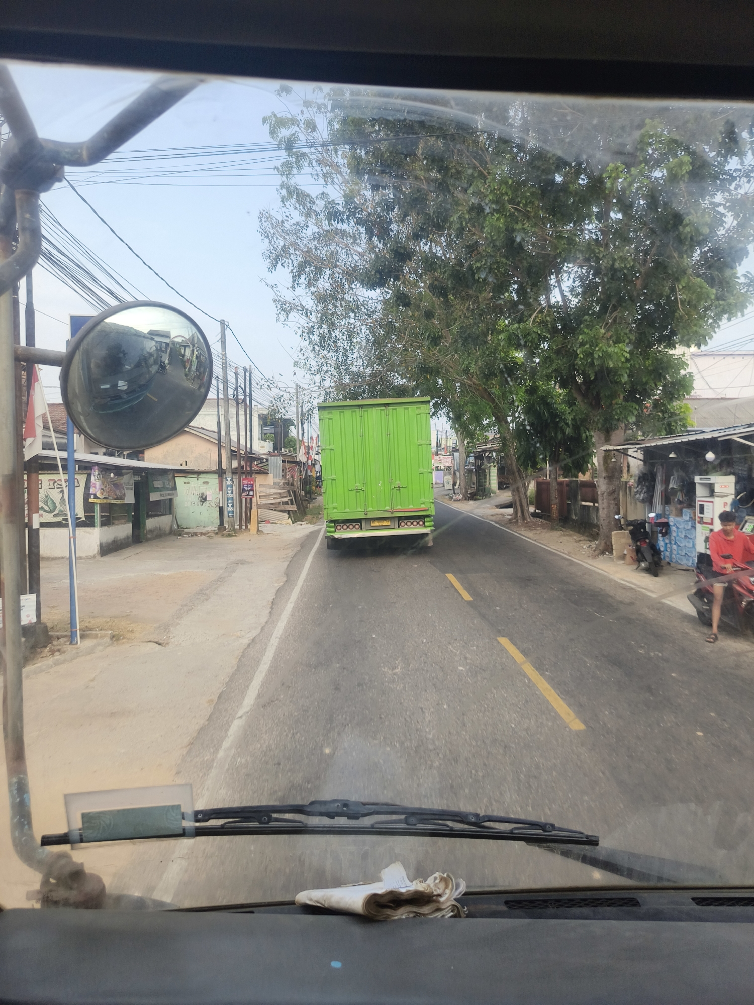 The image shows a view from inside a vehicle, looking forward through the windshield. A bright green truck is driving ahead on a paved road. On the right side of the road, there are several small buildings and stalls, one with a person in a red shirt standing near a motorcycle. There are also trees lining the road and power lines overhead. In the foreground, on the dashboard, there is a folded white cloth. The image appears to be taken in a semi-urban or roadside area in Pangkalpinang, Indonesia, likely during daylight hours.