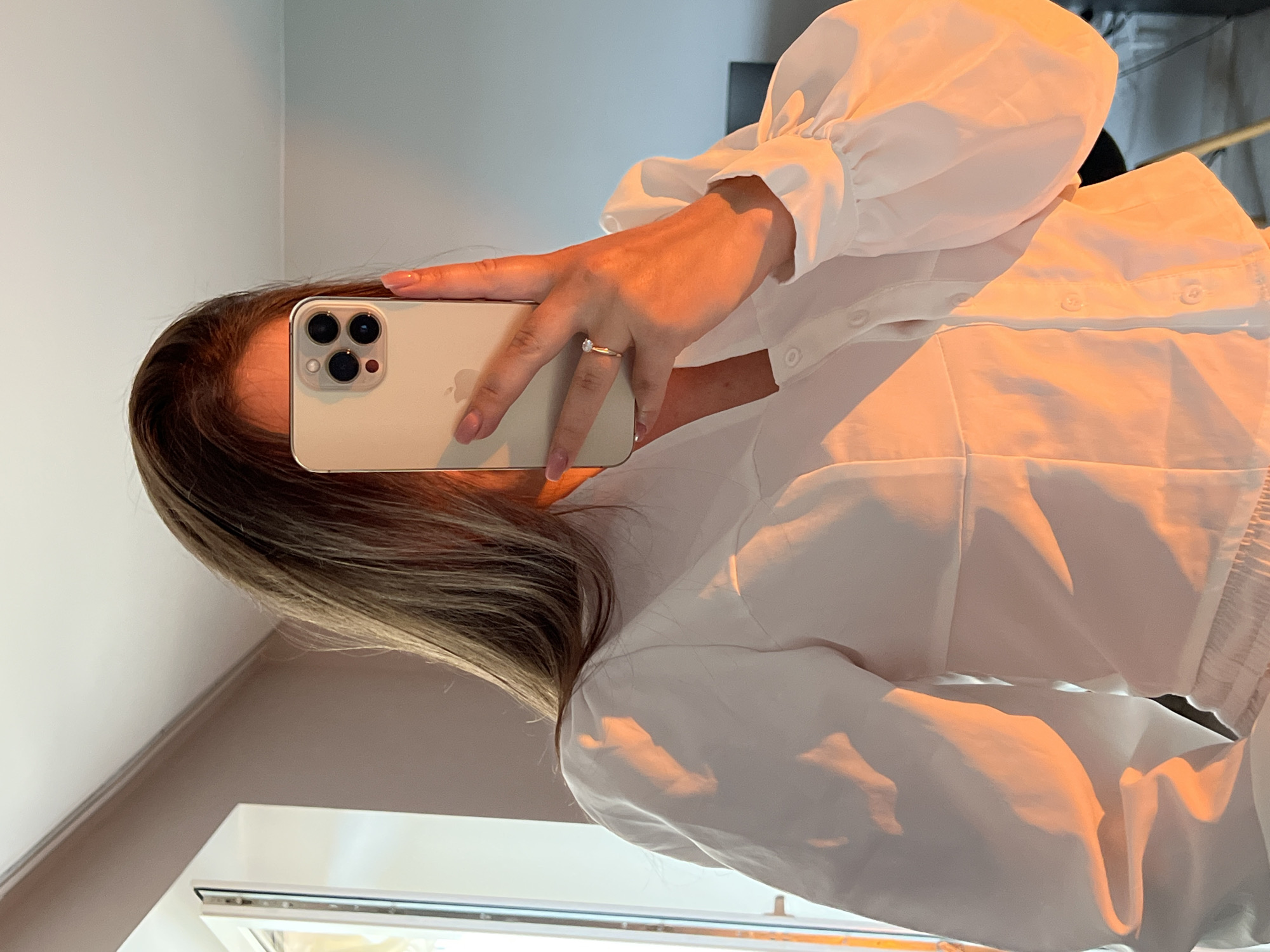 This mirror selfie, rotated 90 degrees counter-clockwise, captures a person taking their own reflection. The subject's face is obscured by a light-colored iPhone, held up to photograph into a mirror. Visible features include long, brown hair with lighter highlights, framing the phone.
The person wears a white, button-down blouse featuring distinctive puffed sleeves with gathered fabric at the cuffs. A delicate ring with a single clear stone adorns the ring finger of the left hand, which also displays manicured nails painted with a light pink polish.
The iPhone, likely a Pro model, prominently displays its three camera lenses on the back. The faint outline of the Apple logo is visible beneath the hand. The indoor setting features light-colored walls and a window frame at the bottom of the image. The scene is illuminated by a warm, golden light, possibly from a sunset or artificial lighting, which creates soft highlights and shadows across the clothing and skin. There are no specific textual elements or visual cues within the image to indicate its location as Ternopil, Ukraine.