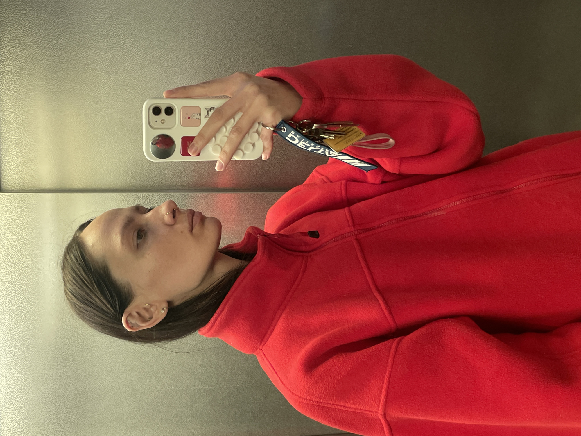A woman takes a selfie in what appears to be an elevator. She is wearing a red fleece jacket and holding a smartphone with a white case decorated with cartoonish images. The phone has keys attached to it via a blue lanyard. The environment is a metal-paneled elevator interior. There is no evidence in the photo that allows for identification of the city, Izhevsk, Russia.