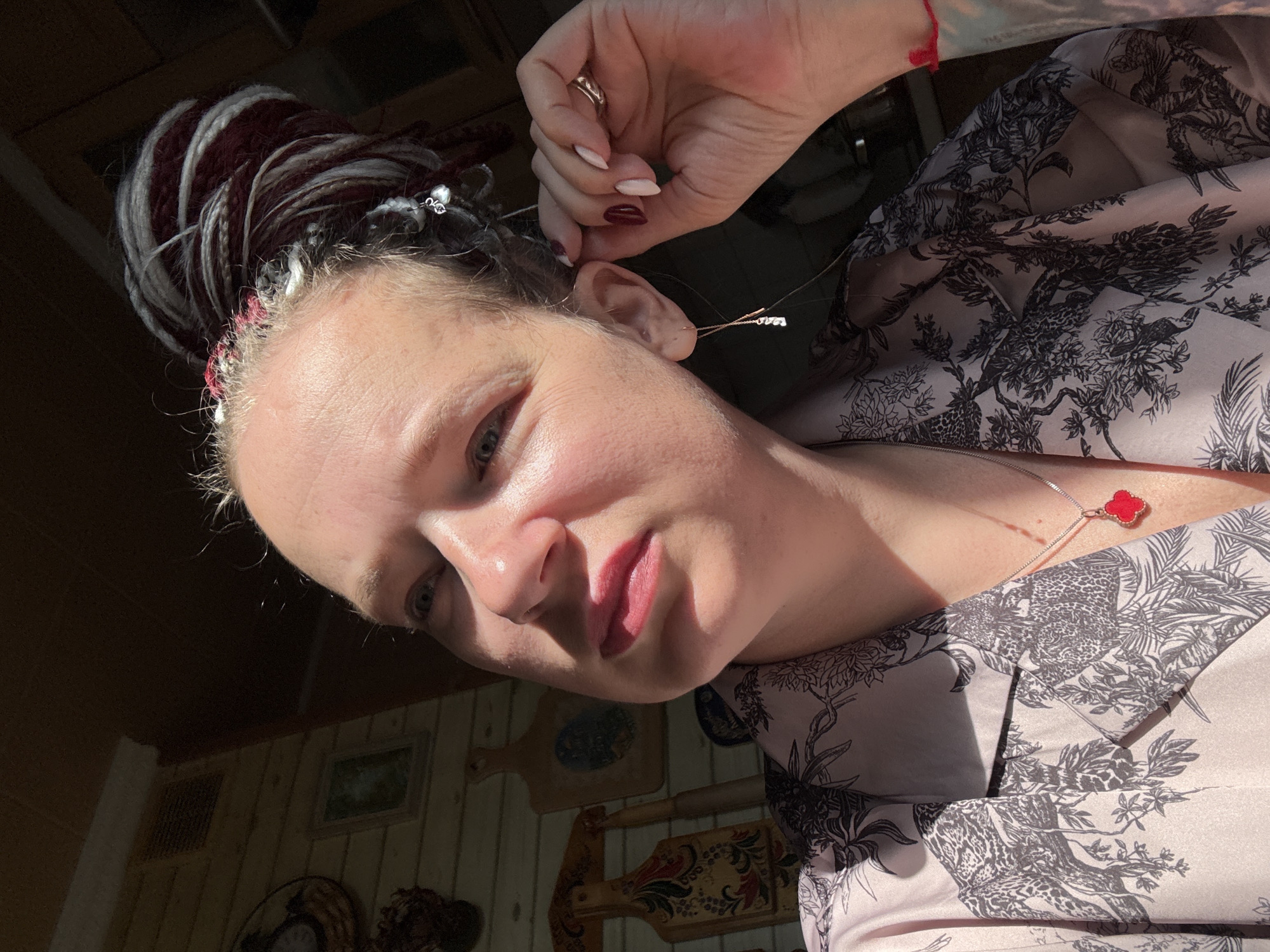 The location depicted is the city of Moscow, Russia.
The image features an adult female, likely in her 30s or 40s, captured in a mid-shot. Her head is tilted slightly upwards, and she is looking towards the upper left. Strong, directional sunlight illuminates the left side of her face (viewer's right), casting sharp shadows across the right side of her face and under her jawline. She has light-colored eyes and her lips are painted with a pinkish-berry shade of lipstick. Her hair consists of thick dreadlocks, a mix of dark red/burgundy and white/grey strands, styled into an updo or bun on top of her head. Small decorative clips or charms are visible within her hair near the top, along with a thin red thread woven near her hairline.
She is wearing a light-colored (pale pink or beige) button-up shirt adorned with a monochrome pattern depicting botanical elements, trees, and what appear to be leopards or other big cats. Around her neck, she wears a silver-colored chain with a bright red, four-leaf clover-shaped pendant. A delicate threader earring with a small white charm hangs from her right ear. Her right hand (viewer's left) is raised, with fingers lightly touching her right earlobe. Her fingernails are manicured, featuring a two-toned polish: light beige/cream on her index and middle fingers, and a dark red/burgundy on her ring and pinky fingers. A silver-colored band ring is visible on her right ring finger.
The background is an indoor setting with horizontal wooden paneling on the walls. Various decorative items are hung on the walls, including several wooden cutting boards painted with folk art designs (e.g., floral patterns, a landscape with a blue lake) and other rustic, dark-toned decorative objects. The upper portion of the background is deeply shadowed, revealing a dark ceiling or corner, with a small vent visible in the upper left.