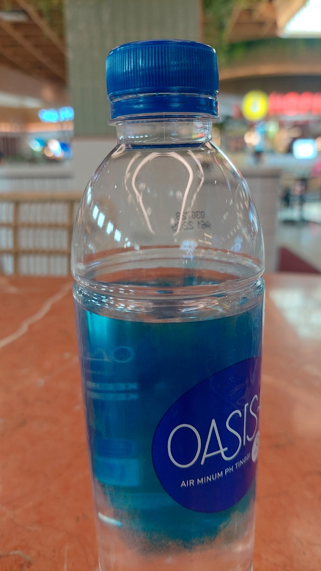 A close-up, eye-level shot shows the top portion of a clear plastic bottle filled with blue liquid, resting on an orange, marbled surface. The bottle has a ribbed blue plastic cap. The label on the bottle is oval, dark blue, with "OASIS" written in white stylized font, followed by smaller white text that reads "AIR MINUM PH TINGGI". The background is blurred, showing the interior of a building, likely a shopping mall, with light-colored architectural elements and colorful out-of-focus lights and signage. The setting suggests an indoor public space in North Jakarta, Indonesia.