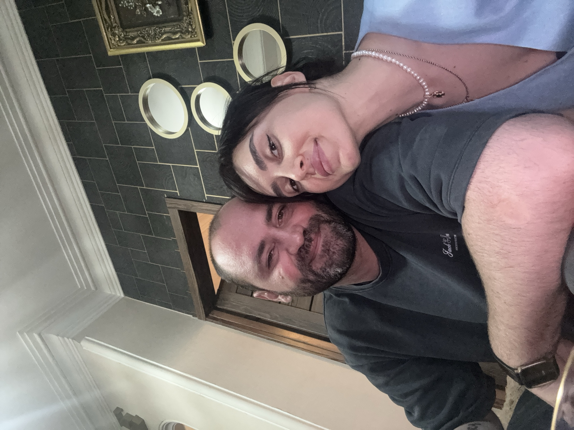 A selfie shows a man and a woman close together. The man has a beard and dark hair, and is wearing a dark gray shirt and a watch. The woman has dark hair, a pearl necklace, and is sticking her tongue out slightly. They are in a room with dark gray tile walls decorated with small gold-framed mirrors and a gold-framed painting. The location may be in Tbilisi, Georgia, but this cannot be confirmed from the image alone.