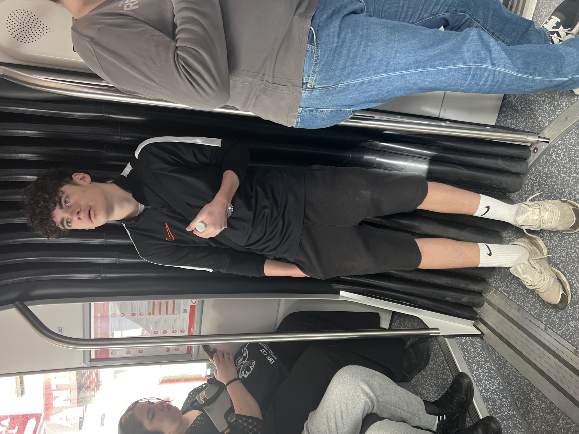 The image shows the interior of a tram in Strasbourg, France. Several people are visible. In the foreground, a person in grey sweatpants and black shoes sits next to someone wearing a black t-shirt and reading a mobile phone. Behind them, a young man in a black tracksuit with a red Nike logo on the jacket and white socks is lying across the seats. Above him, another person is standing, wearing blue jeans and a grey long-sleeved shirt. The background reveals advertising signage and windows. The floor is a grey speckled material. The seats are black.