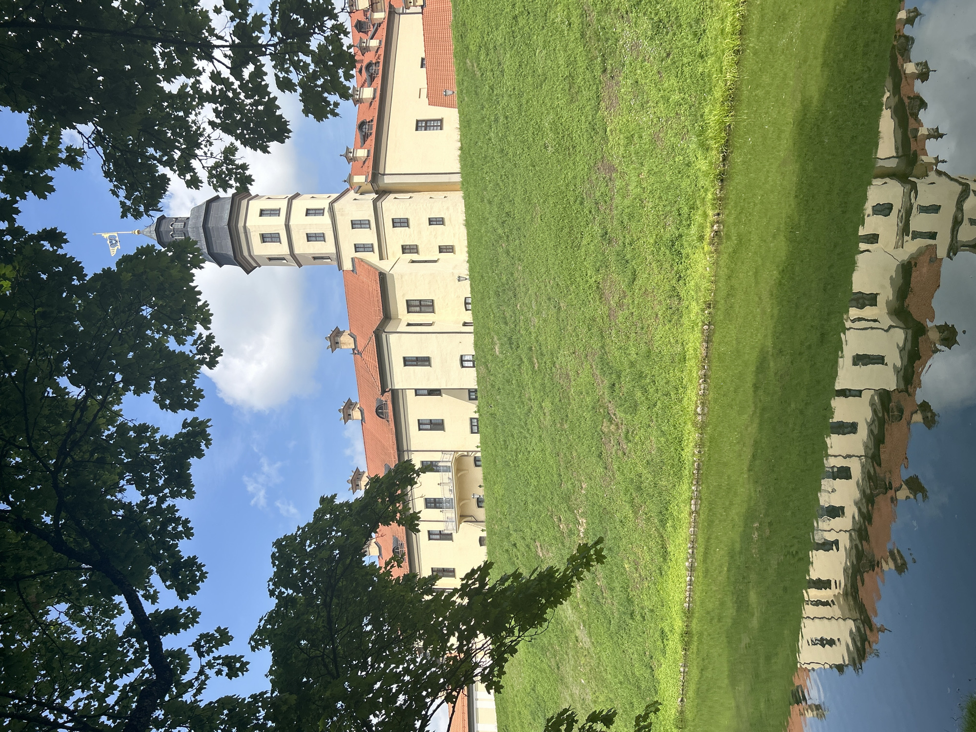 This is a medium shot, taken at eye level, of the Nesvizh Castle in Nesvizh, Belarus. The castle is a large, yellow building with a red tile roof, situated on top of a grassy hill. A tower with a dark roof and a flag on top stands prominently on the right side of the castle. The castle is reflected in the calm water of a moat in the foreground. Lush green trees with dark leaves frame the scene from the top and left. The sky is blue with scattered white clouds. There are no people visible in the image. The overall scene is serene and picturesque, showcasing the architectural beauty of the castle and its natural surroundings.