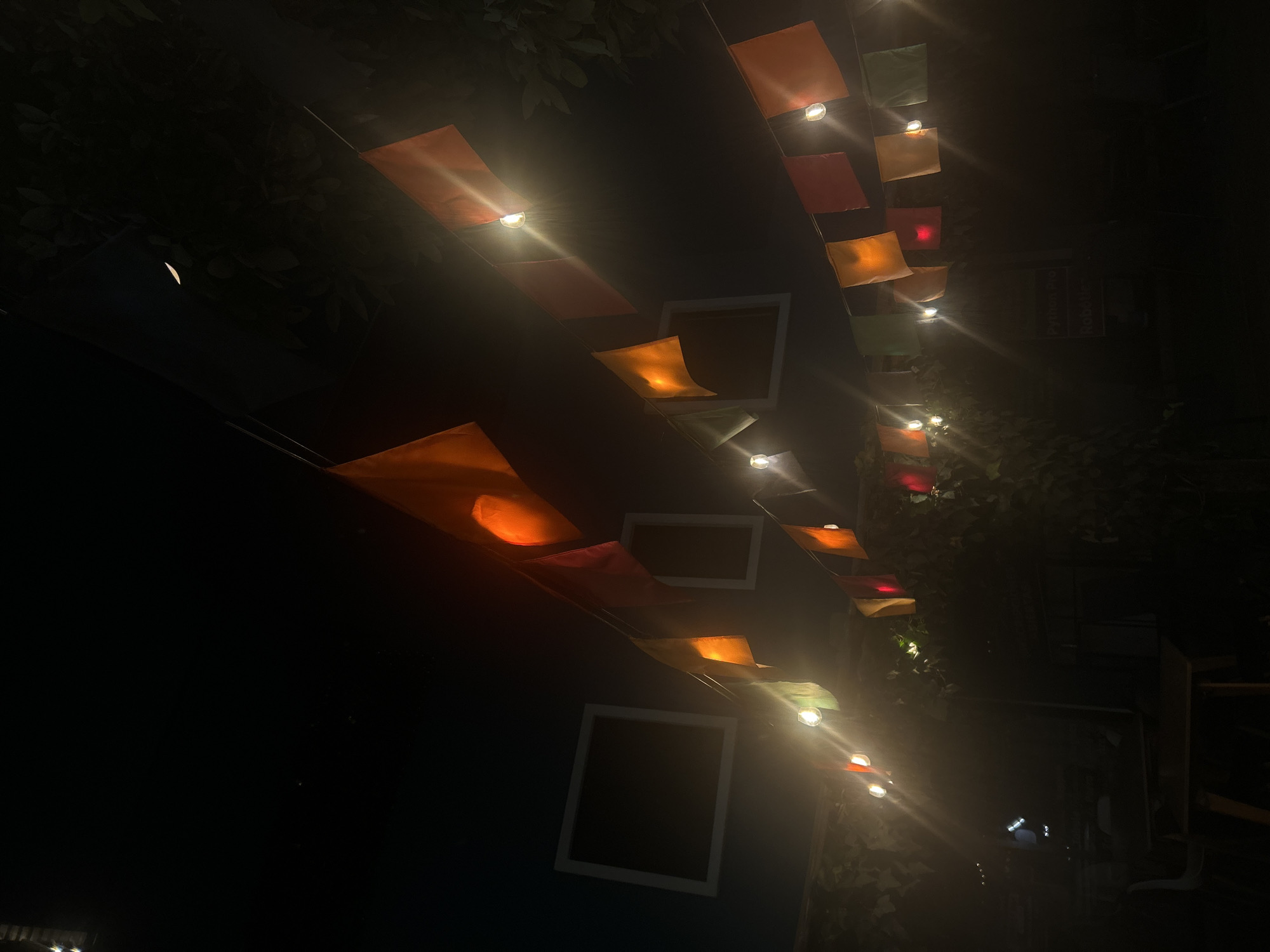 The image captures a dimly lit outdoor setting at night, likely a patio or backyard in Unknown, Unknown. Strings of colorful, square-shaped lanterns are suspended overhead, each illuminated by a single light bulb. The lanterns vary in color, including shades of orange, red, yellow, and green. The light from the bulbs casts bright flares, while the rest of the scene is mostly in shadow. Foliage, possibly trees or climbing plants, frames the lanterns, and a structure resembling a building is visible in the background. Some of the lanterns are encased in square frames. The photograph has a slightly tilted perspective, looking upwards at the lanterns.