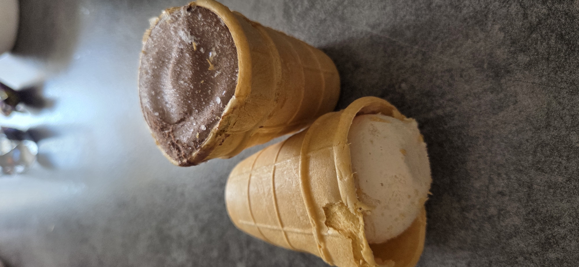 Two ice cream cones are placed on a dark grey, textured surface. One cone, on the right, is filled with chocolate ice cream that has a slightly rough, crystalline texture on top, possibly due to freezing. This cone is upright. The second cone, on the left, is lying on its side, with the opening facing upwards. It contains a pale, creamy-colored filling, which appears to be a type of ice cream or mousse. The waffle cones are golden brown and have a grid pattern. The background is blurred, showing hints of light sources and possibly other objects, but these are not clearly identifiable. The location is not specified in the image.