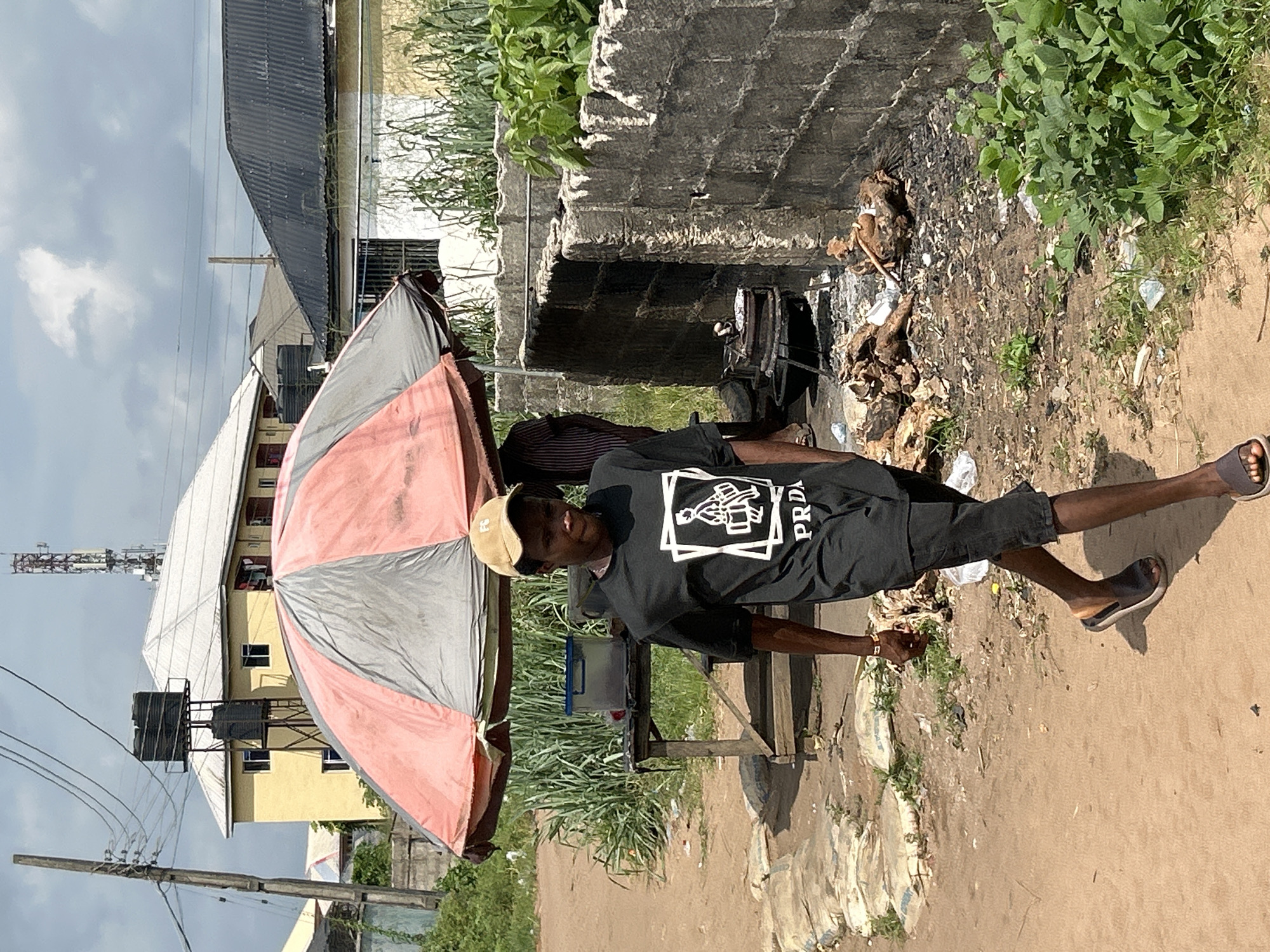 The image shows a man standing in what appears to be a street or roadside environment in Ihiagwa, Nigeria. He is wearing a black t-shirt with a design on it and the word "PRDA" written on the side, black shorts, a tan cap, and sandals. He stands next to a makeshift table or stand sheltered by a large, two-toned umbrella, likely offering shade from the sun. Behind him is an open drainage or concrete channel, with a visible accumulation of trash and debris. The surrounding area suggests an urban or semi-urban setting with buildings, including one with water tanks on the roof, and overgrown vegetation. Power lines and a tower are also visible in the background, contributing to the urban landscape.