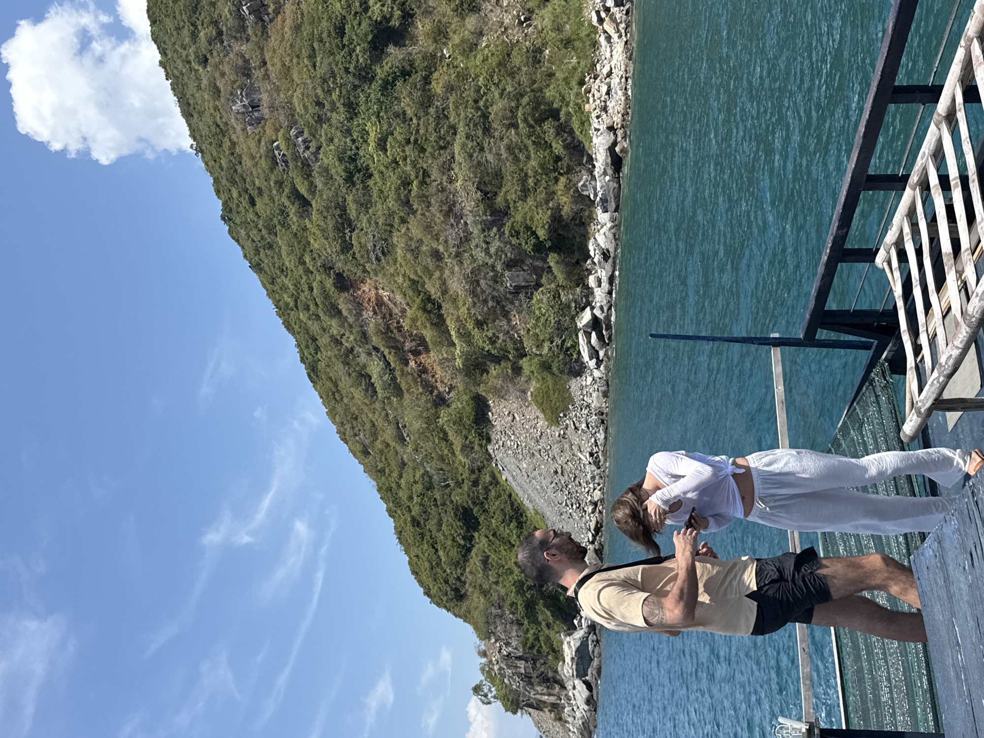 A man and a woman stand on the deck of a boat, with the deck railing in the lower right corner. The man wears a tan shirt and black shorts, while the woman wears a white shirt and pants. The boat is on the water, with a green, rocky cliff in the background. The sky is blue with some white clouds. The scene appears to be near Bac Nha Trang Ward, Vietnam, given the landscape features.