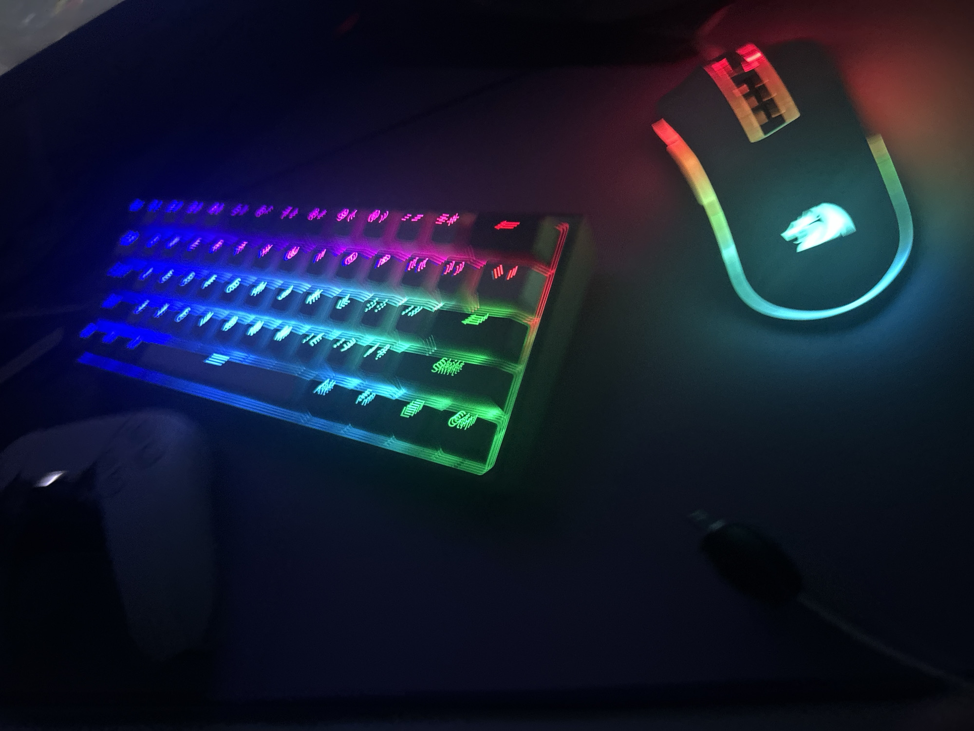 In a dimly lit room, a gaming keyboard with vibrant RGB lighting and a gaming mouse with similar illumination are laid out on a dark surface, likely a desk. The keyboard is positioned diagonally from the bottom left to the top right of the frame, with its keys displaying a gradient of colors from green to blue and purple. The mouse is located in the lower right portion of the image, its shape outlined by a luminous ring of colors that shift from blue to red. A black cable extends from the left side of the frame, presumably connected to the keyboard. In the upper left corner, a blurred object, possibly a white gaming controller or headset, is partially visible. The scene appears to be indoors and is not indicative of any specific location within the city of Shumen, Bulgaria, as no contextual elements or identifying features of the city are present in the image.