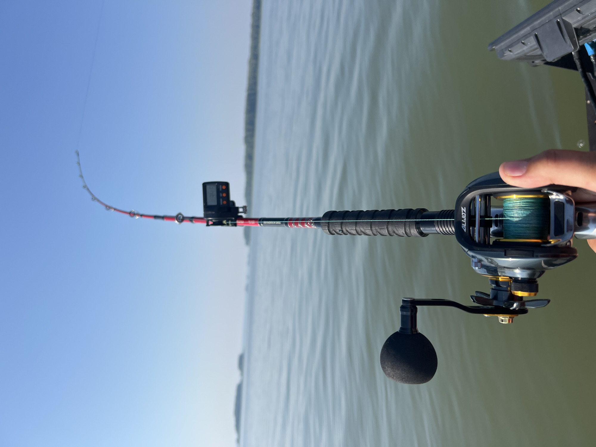 An over-the-shoulder shot shows a person's hand holding a fishing rod and reel. The rod is red and black with a digital counter attached. The reel is black and gold, with green fishing line. The rod is extended over a body of water, which is a calm green with a few small islands in the distance. The sky is clear blue. The scene appears to be from a boat, as a black electronic device is visible on the right side of the frame. The location is the city of Novopichugovo, Russia.