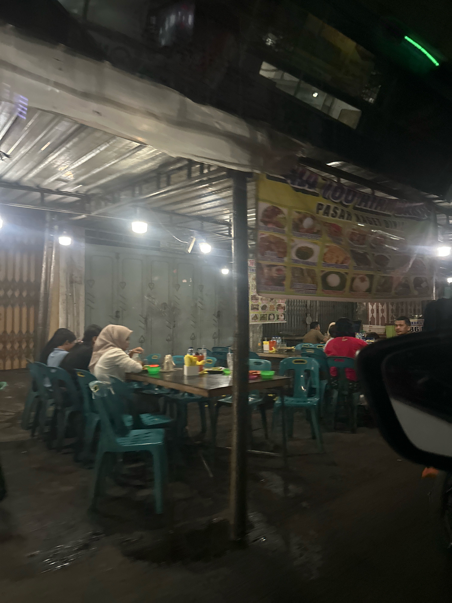 The image shows an outdoor dining area at night, likely in the city of Binjai, Indonesia. Several people are seated at tables, some of whom are eating. The scene is illuminated by overhead lights. A banner with images of food is displayed above the tables. The ground appears wet, possibly from rain. The overall atmosphere is casual and appears to be a local eatery.