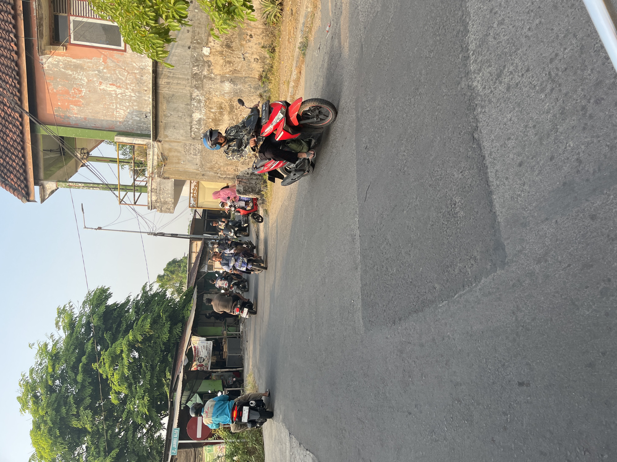 The image captures a street scene in Tenggulunan, Indonesia, on a sunny day. The road surface is asphalt, showing signs of wear with some patching. On the left side of the road, there are several small shops or stalls with corrugated metal roofs, and a large tree with dense green foliage. A street sign is visible, reading "Jl. Jambu Alas".
Multiple people are riding motorcycles, moving in both directions on the road. One person in a bright blue jacket and black pants is riding a black motorcycle away from the camera on the left. Further down the street, a man in a brown shirt is on a scooter. A man in a blue and white shirt is on a blue motorcycle, and several other motorcycles are also visible in the middle ground.
On the right side of the road, a building with a weathered, peeling orange and pink facade stands. It has a balcony with a green railing and a red tile roof. A man wearing a camouflage-patterned jacket and a blue helmet is riding a red motorcycle towards the camera in the foreground. He is wearing black pants and flip-flops. Another motorcycle with a woman wearing a pink headscarf and a child is parked nearby. The background shows a clear sky and more buildings and trees. The overall impression is of a typical day in a local Indonesian community with active street life centered around motorcycle transportation.