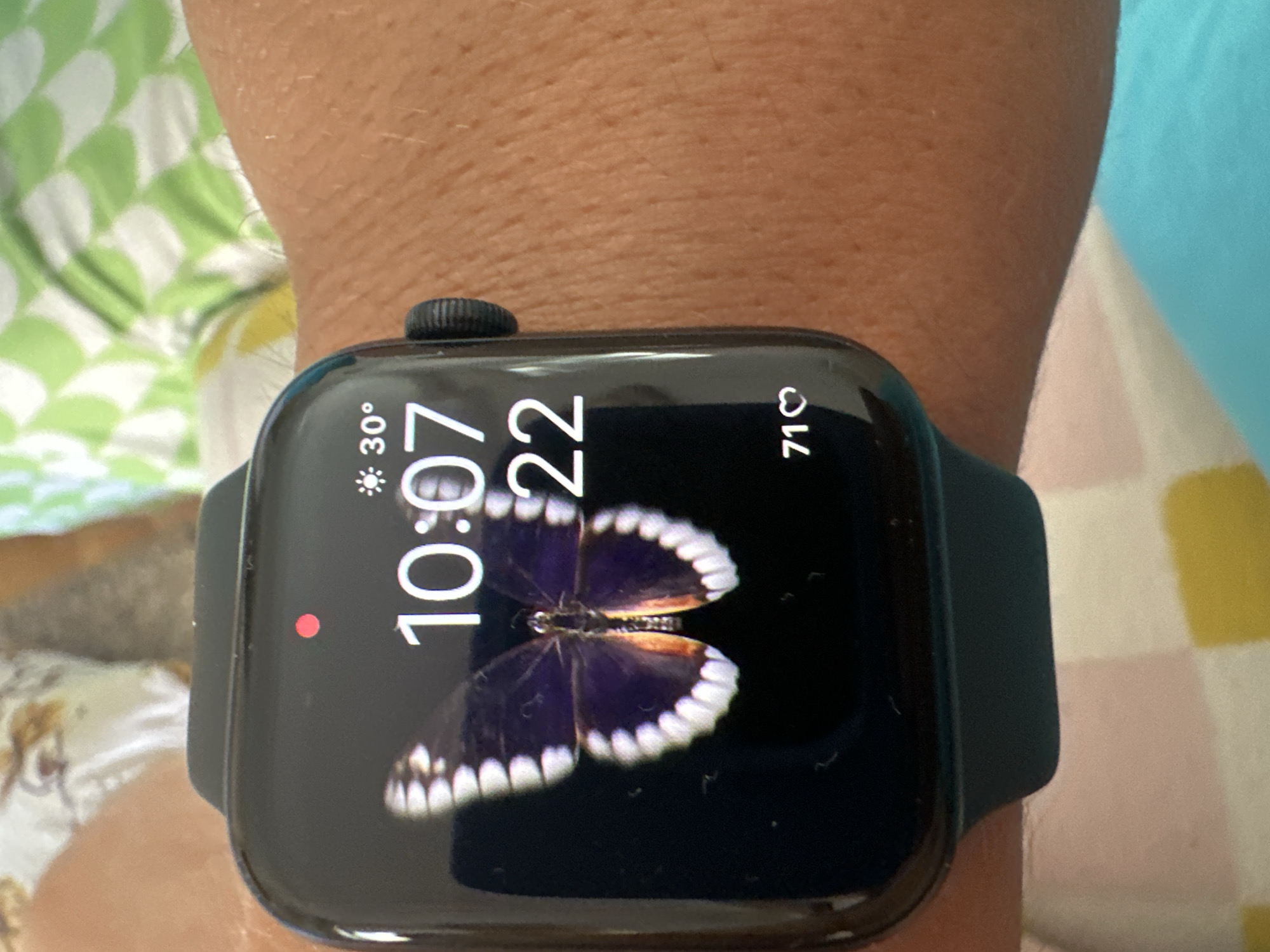 The image shows a close-up of a dark smartwatch worn on a person's wrist. The smartwatch screen displays the time as 10:07 and 22. There is also a weather icon indicating 30 degrees, and a heart icon with the number 71, likely representing a heart rate. A colorful butterfly graphic is displayed as the watch face. The background is blurred but appears to be a patterned fabric. The image does not contain any information about the city of Budva, Montenegro.