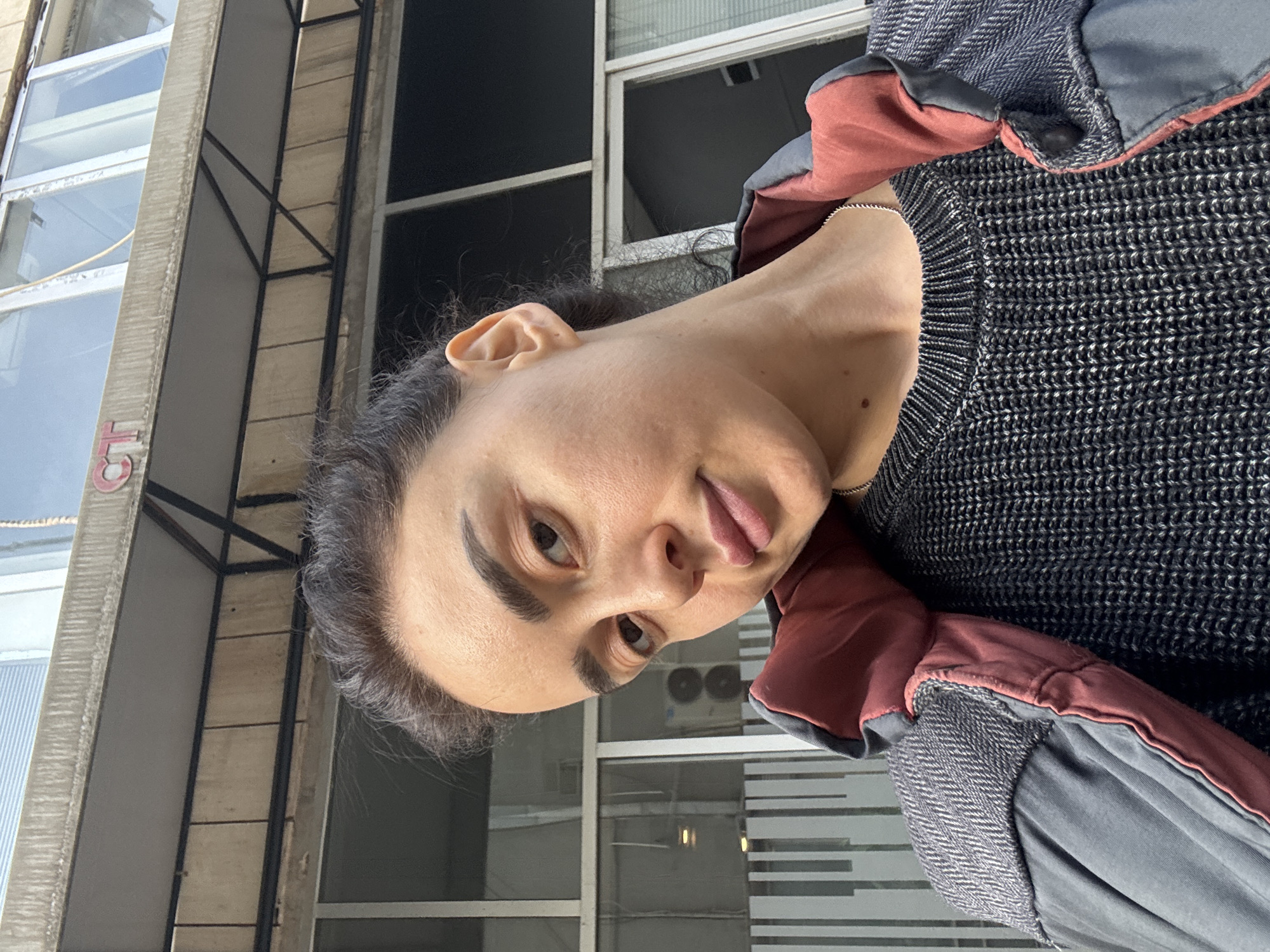 A medium close-up, portrait-oriented image, rotated 90 degrees counter-clockwise, shows a person looking directly at the camera with a playful expression, their tongue slightly protruding. The individual has dark hair and eyebrows, and an olive skin tone. They are wearing a dark, textured knit sweater and a layered jacket; the inner lining of the jacket is a reddish-brown or terracotta color, while the outer layer is greyish-blue with a herringbone pattern visible on the sleeve. A thin silver chain is draped around their neck.
The background depicts an urban environment, identified as Tbilisi, Georgia, featuring tall buildings with multiple windows. Some windows reflect bright daylight, suggesting a sunny or clear day, while others appear dark. Visible architectural elements include concrete pillars or beams and dark metal frames. In the upper left background, on a concrete structure, partially visible red letters resembling "CT" or "GT" can be seen. An external air conditioning unit is also discernible on a building in the mid-ground. The overall setting is an outdoor, daytime urban scene.