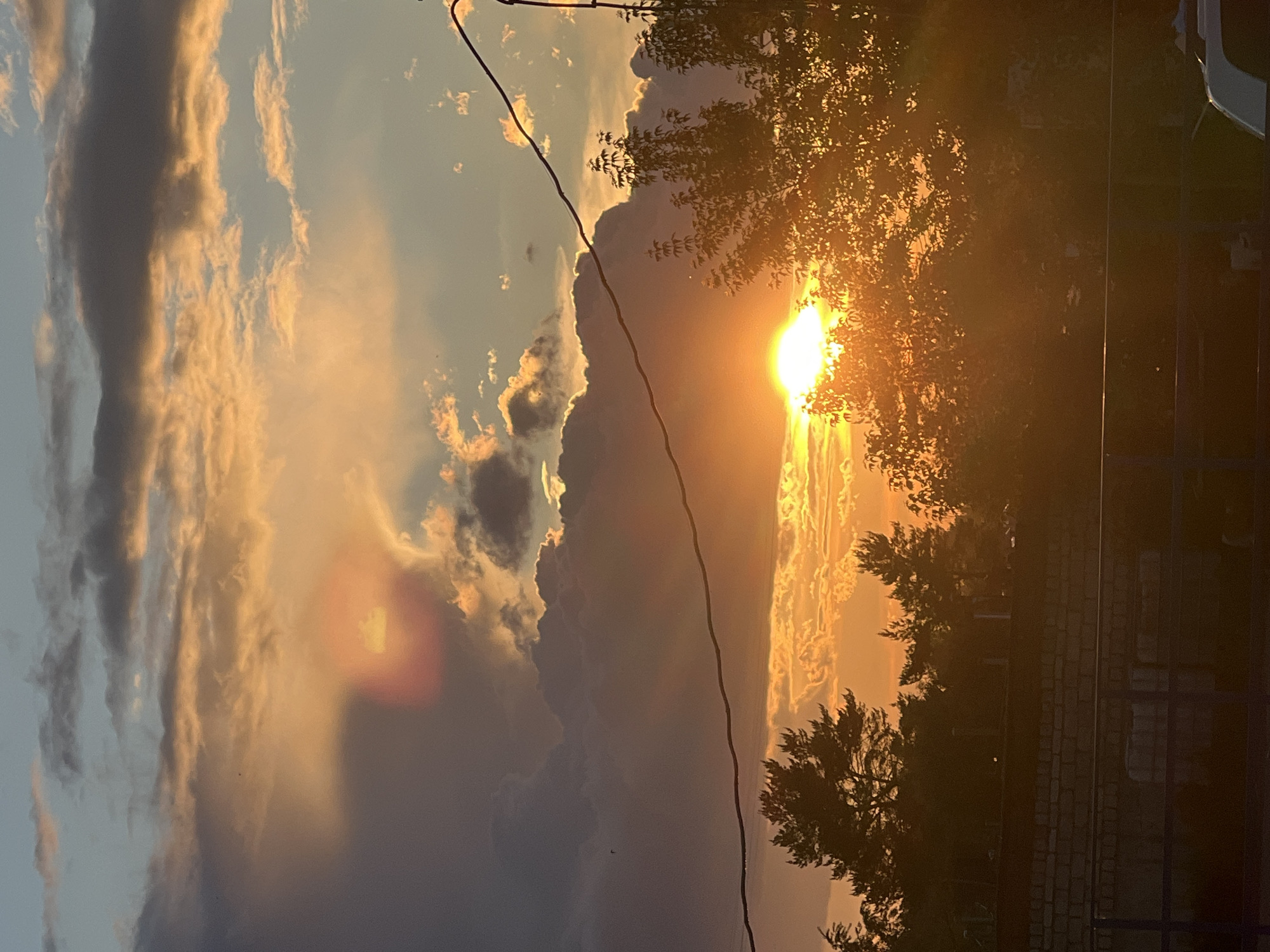 Captured at sunset in Chaikovsky, Russia, the photograph displays a partially obscured sun radiating golden light through scattered clouds and tree silhouettes. A power line cuts diagonally across the frame. A brick structure is visible along the lower right corner.