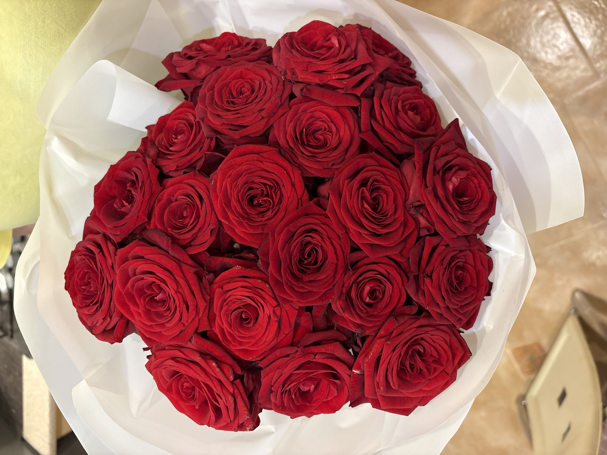A bouquet of approximately 20 red roses is presented, wrapped in white paper. The roses are in full bloom, with deep red petals and tightly packed centers. The wrapping paper is a bright white and appears to be a matte finish, with a slightly crinkled texture in places. The bouquet is held upright, suggesting it has been recently arranged or is being presented. The background is out of focus, but hints of a warm-toned floor and a blurred object with a reflective surface are visible. The overall impression is one of a romantic gesture or a decorative centerpiece. The image is shot from a slightly elevated angle, looking down at the bouquet. There is no indication of the city of Minsk, Belarus, or any specific location.