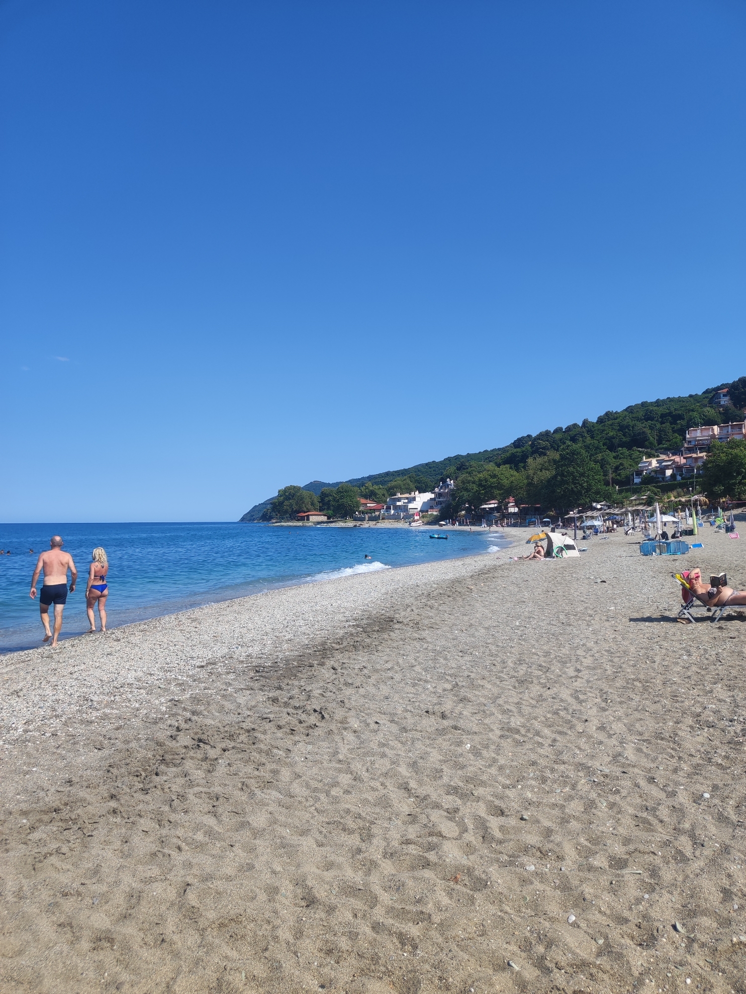 The image shows a beach in the Municipal Unit of Melivia, Greece. In the foreground, a man and a woman are walking on the sandy beach, approaching the water. The man is wearing dark swim trunks, and the woman is wearing a blue bikini. The beach has a transition from sand to pebbles closer to the water. In the background, the turquoise sea meets the shore, and beyond that, a tree-covered hillside is dotted with white buildings. Further down the beach, several people are seen relaxing under umbrellas, sunbathing on lounge chairs, or swimming. The sky is clear and bright blue.