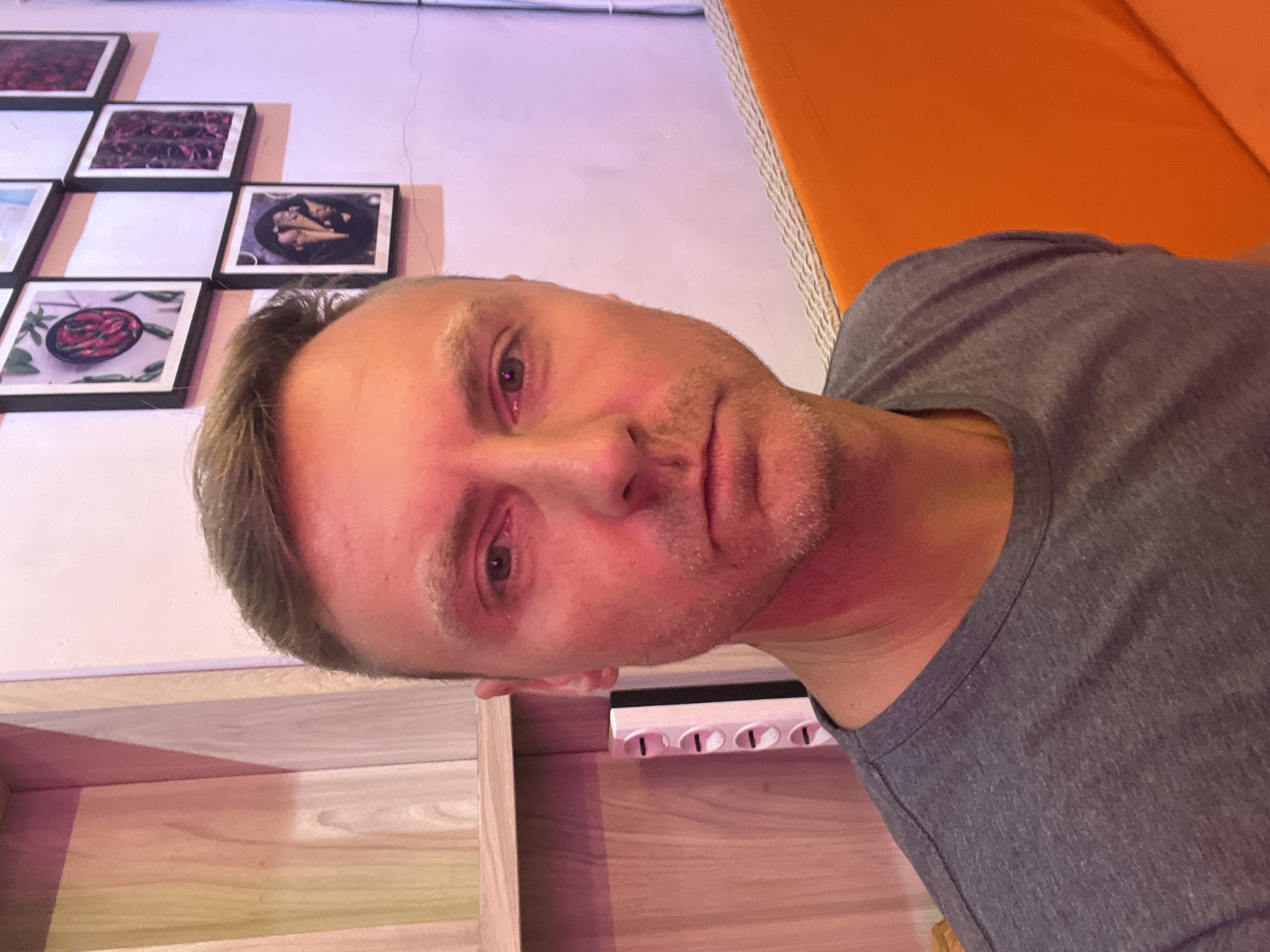 This indoor portrait features a man looking directly at the camera, likely a selfie. He has fair skin with a light stubble, short light-colored hair, and is wearing a grey t-shirt. His expression is neutral, and his eyes appear somewhat red or irritated.
The background reveals a wall decorated with multiple framed square pictures arranged in a grid. These pictures depict various food items, including red chili peppers in a black bowl and what appears to be cooked meat or poultry. This suggests a setting like a restaurant or cafe. To the man's right, a section of an orange upholstered seat or cushion is visible. Below him, there is light-colored wooden furniture, possibly a shelf or table, on which a white power strip with four outlets is placed.
The scene is illuminated by a prominent pinkish-red ambient light, casting a distinct hue over the man's face, neck, and the wall. The image provides no discernible text. The location context is Sukawati, Indonesia.