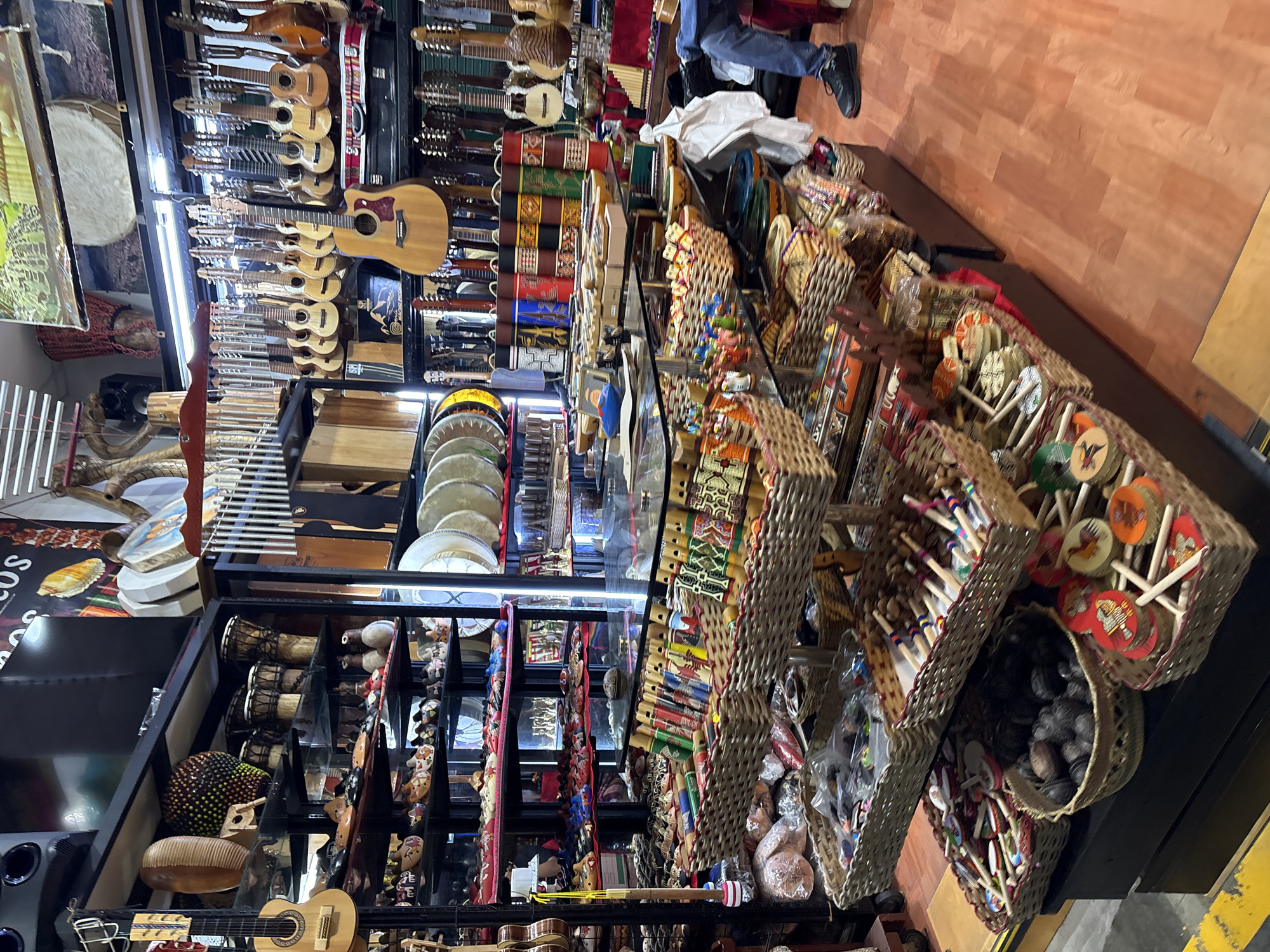 The image shows the interior of a shop filled with handicrafts and musical instruments. In the foreground, several woven baskets are stacked, containing an assortment of colorful items. Some baskets hold what appear to be small wooden toys with spinning tops, while others contain dark, round objects that could be gourds or seed pods. Further back, more woven baskets overflow with intricately decorated flutes or panpipes.
On shelves behind the baskets, a variety of items are displayed. These include what look like decorative clay figures, intricately woven textiles, and more musical instruments such as drums and stringed instruments. The walls are lined with shelves holding numerous guitars, ukuleles, and what appear to be other types of stringed instruments, many of them hanging. Some shelves also display larger percussion instruments like drums and what might be a marimba or xylophone.
The shop has a warm, wooden floor. In the lower right corner of the image, a person wearing blue jeans and a black shoe is partially visible, suggesting a customer or employee. The lighting appears to be a mix of overhead shop lights and natural light, creating a well-lit but also somewhat cluttered atmosphere. The overall impression is a vibrant and eclectic market stall or shop.
This scene is consistent with the types of artisan shops found in Miraflores, Peru, particularly in areas known for their craft markets like the Mercado Indio.