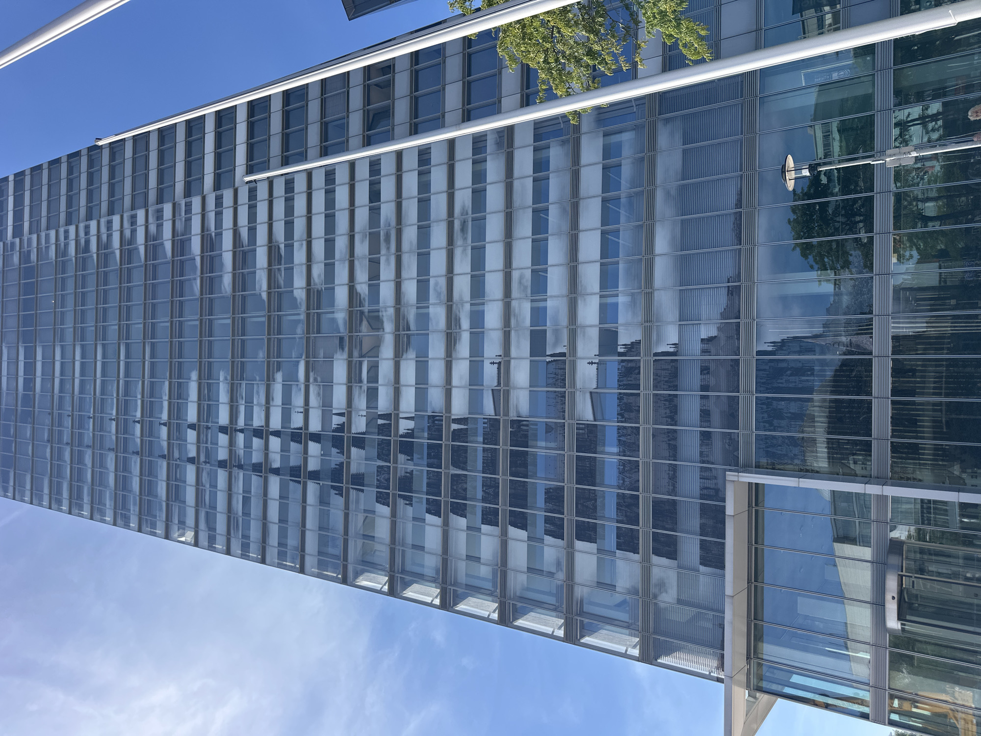 The image shows a contemporary glass skyscraper in Cologne, Germany, with a prominent reflection of Cologne Cathedral on its facade. The skyscraper, likely the Kölnturm, is viewed from a low angle, emphasizing its height against a clear blue sky with scattered white clouds. The glass exterior reflects the surrounding environment, most notably the intricate spires and Gothic architecture of the cathedral, creating a striking contrast between modern and historic structures.
At the base of the skyscraper, there is an entrance area with a revolving glass door and a modern awning. Two tall, white poles are visible on the right side of the frame, extending upwards parallel to the building. Green trees are also visible to the right of the building, with their leaves catching the sunlight. The overall scene suggests a bright, sunny day in an urban setting.