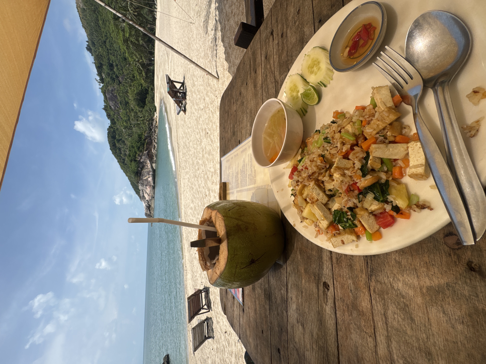 The image captures a meal at a seaside restaurant in Baan Thong Nang, Thailand. The scene is set on a wooden table overlooking a sandy beach and the ocean. A plate of fried rice with tofu and vegetables is the main focus, accompanied by side dishes of cucumber slices, a lime wedge, and two small bowls of sauce. Eating utensils (fork and spoon) are also present on the plate. A fresh coconut with a straw rests next to the plate. In the background, the beach stretches out towards the ocean, and lounge chairs are visible on the sand. A lush, green hillside rises in the distance, creating a scenic backdrop. The sky is partly cloudy.