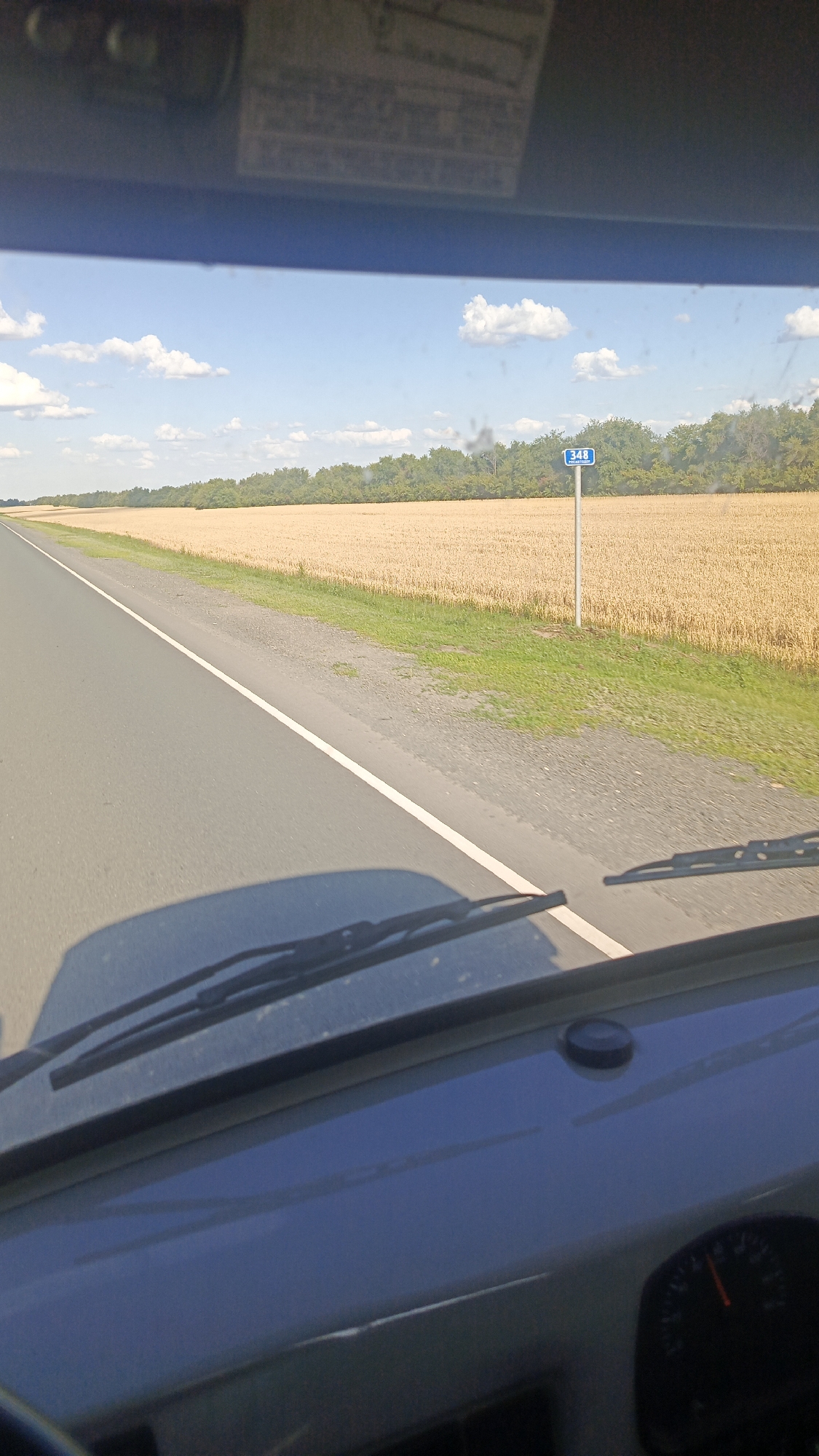 The image shows a view from inside a vehicle, looking out through the windshield at a rural landscape. A paved road stretches into the distance, bordered by a field of golden wheat on the right. A line of green trees forms the horizon beyond the field. A blue sign with the number "348" is visible on a post beside the road. The sky is blue with scattered white clouds. The dashboard of the vehicle is visible in the foreground, with the windshield wipers resting on the glass. The scene appears to be a sunny day in the countryside, possibly on a highway or rural road. The location is identified as Zolotaya Step, Russia.