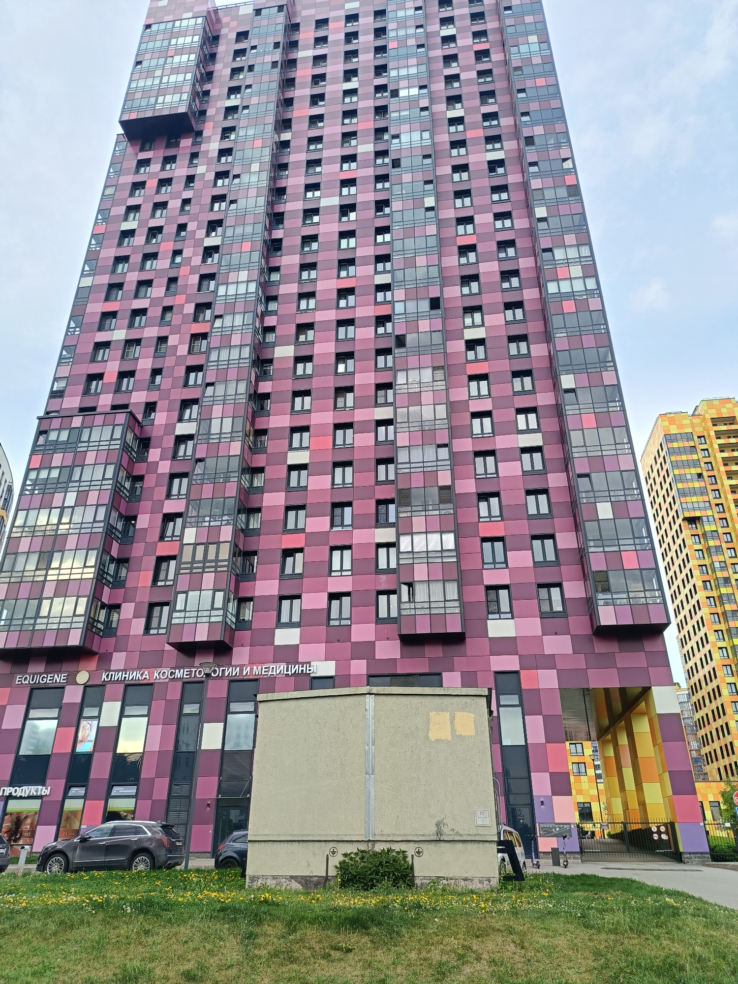 The image depicts a tall, modern residential building in Saint Petersburg, Russia. The building's facade is characterized by a patchwork of various shades of pink and purple, creating a checkered pattern. The building features numerous windows, and several sections protrude outward, forming balconies or extended living spaces.
A shorter, yellow-and-orange building is visible in the background to the right. In the foreground, there is a green lawn with dandelions. A utility box sits on the grass near the building. To the left, a parked car is visible in front of commercial storefronts or offices. The storefronts have signage in Russian, including the word "КЛИНИКА", suggesting a clinic. A person walks along the sidewalk on the right side of the image. The sky is overcast.