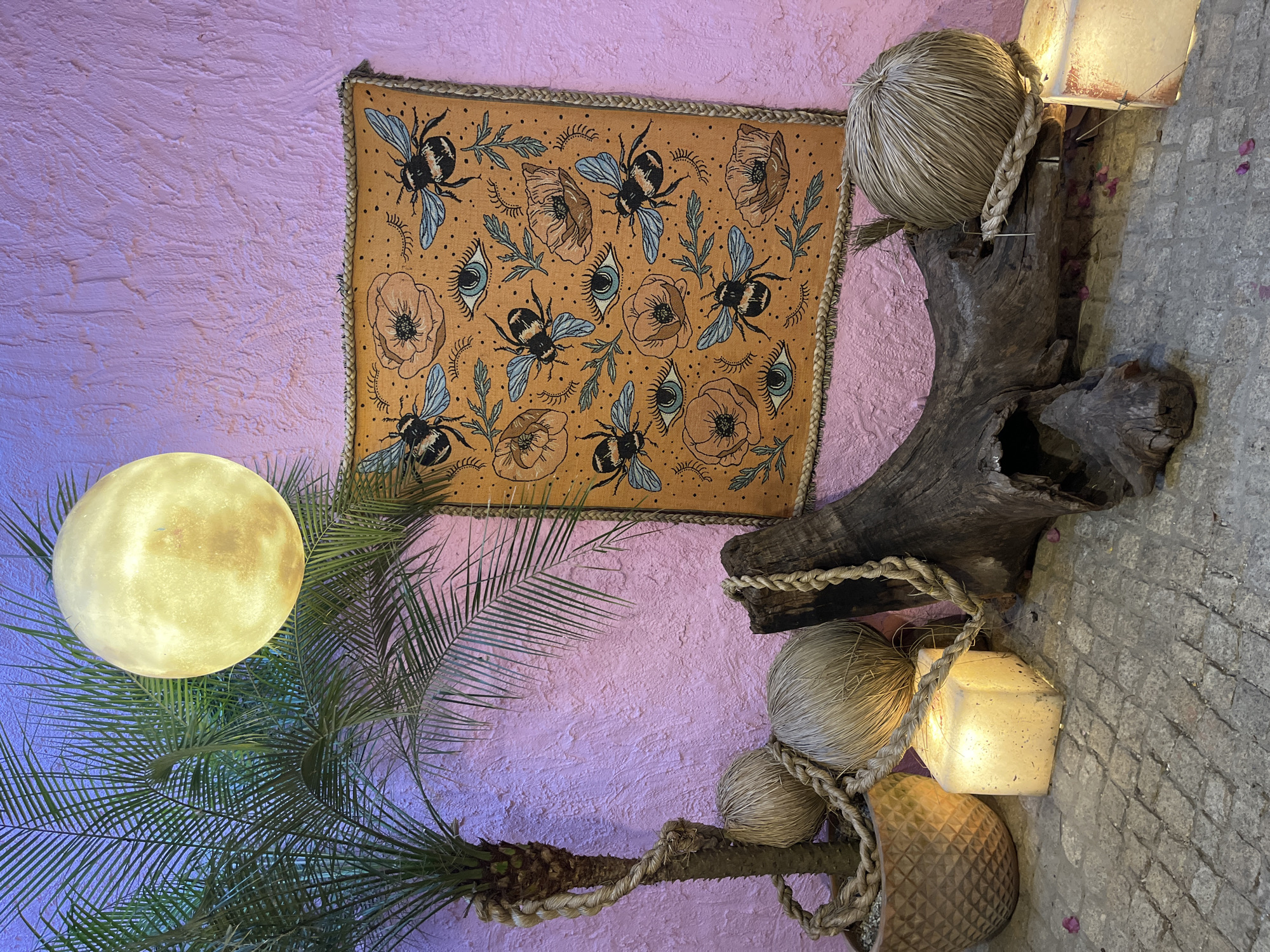 The image presents an interior display against a lavender-textured wall. A square textile featuring a repeating pattern of bees, flowers, and eyes is hung on the wall. To the right, a gnarled piece of driftwood is adorned with woven orbs and a cube-shaped lamp, all connected by rope. A palm tree with a spherical light fixture stands to the left. The wall is of indeterminate interior design. The flooring or adjacent surface is of a gray, brick-like material. The location is Unknown, Unknown. No people are visible.